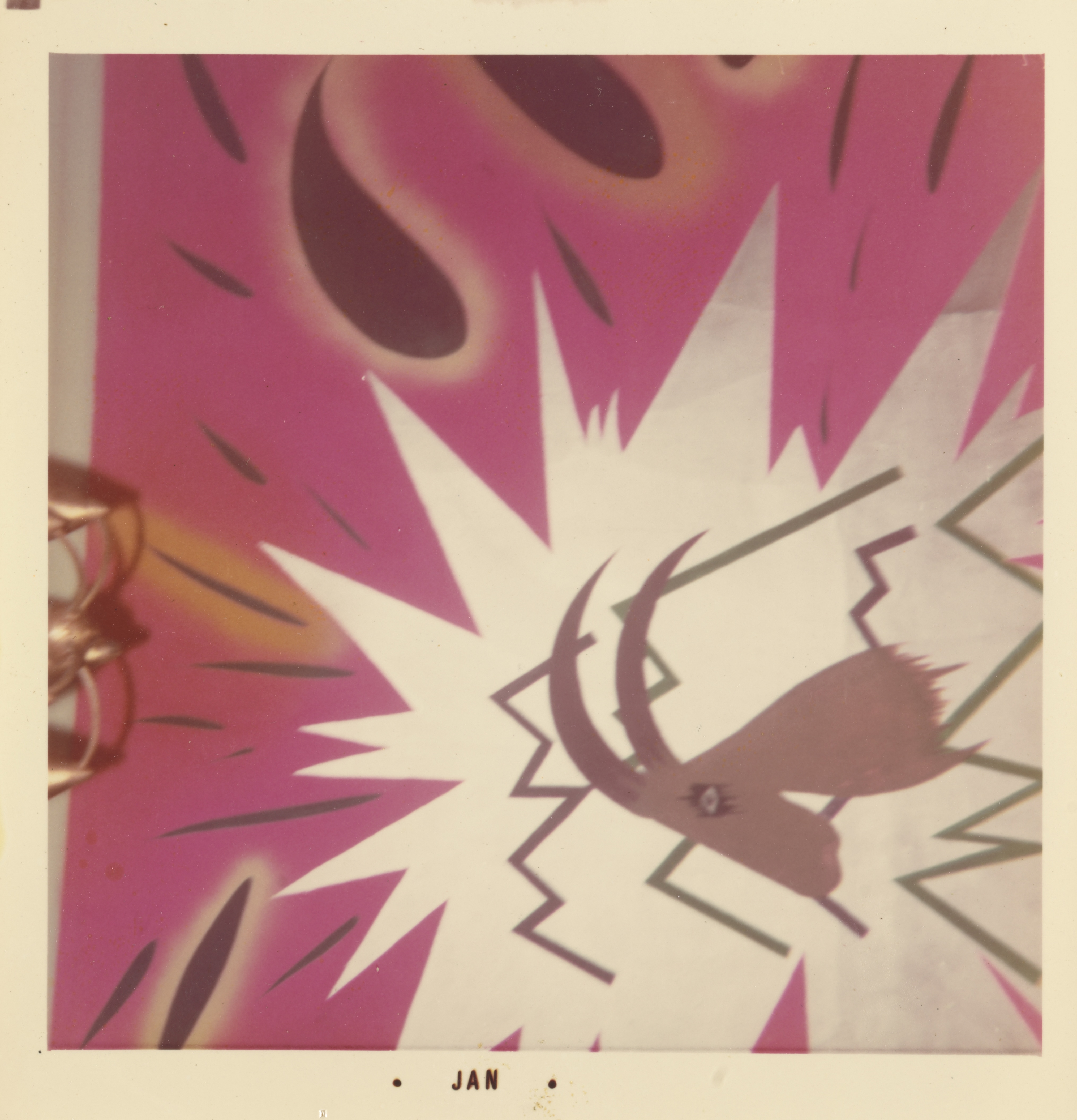 Photograph of a bold, graphic mural with a hot pink background and jagged white burst at the center. In the foreground, a stylized brown goat head with large curved horns and a single visible eye is framed by sharp black and green zigzag lines. Teardrop shapes and angled black marks are scattered across the upper portion. The white border of the print includes the text “• JAN •” centered along the bottom edge.
