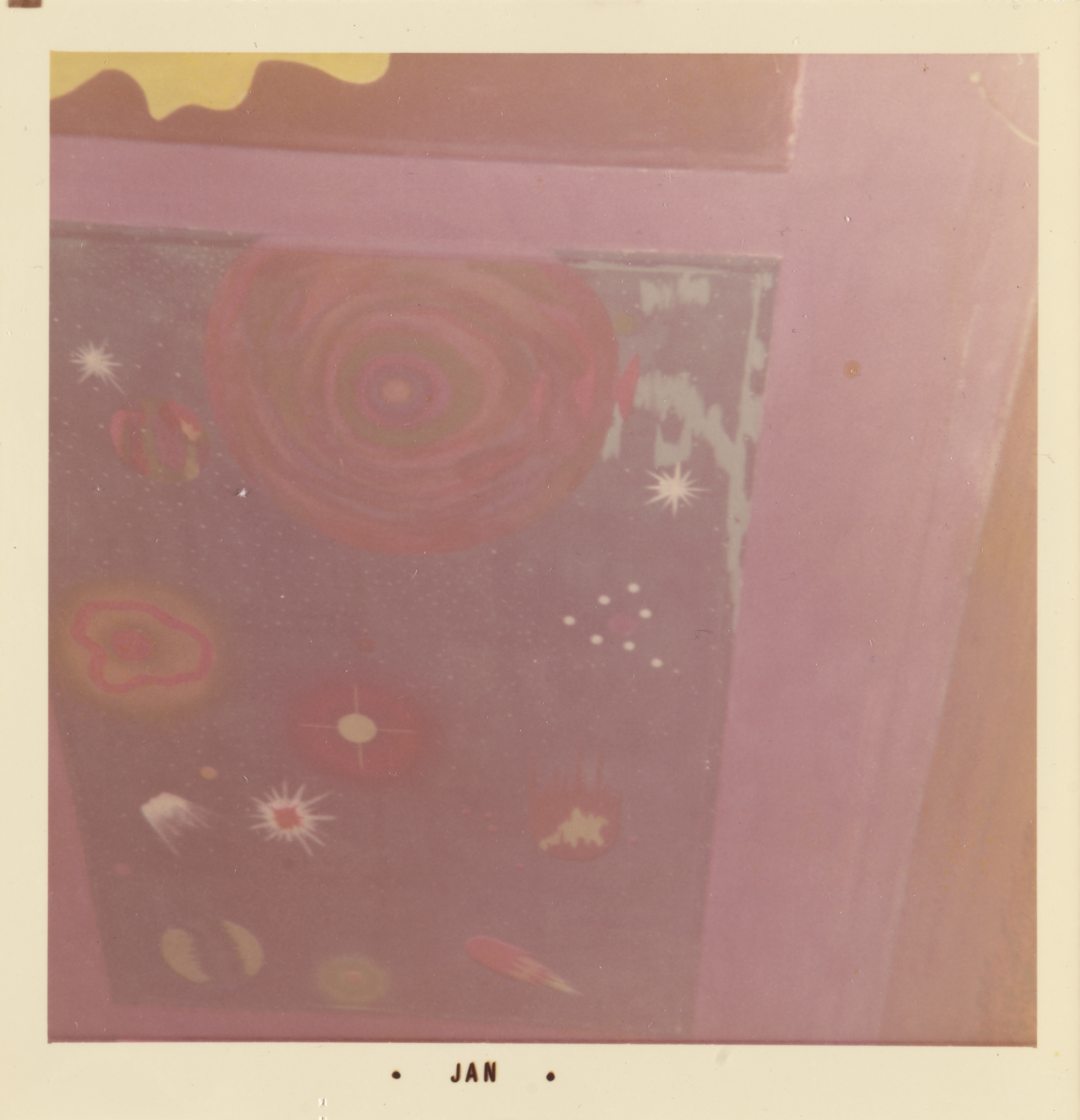 Photograph of a painted panel featuring a cosmic scene with stylized celestial forms. A large, concentric red and green circle dominates the upper left, surrounded by various abstract planets, stars, and comet-like shapes in red, orange, white, and green. The panel is bordered in pink, with part of a yellow organic shape visible at the top. The white border of the print includes the text “• JAN •” centered along the bottom edge.