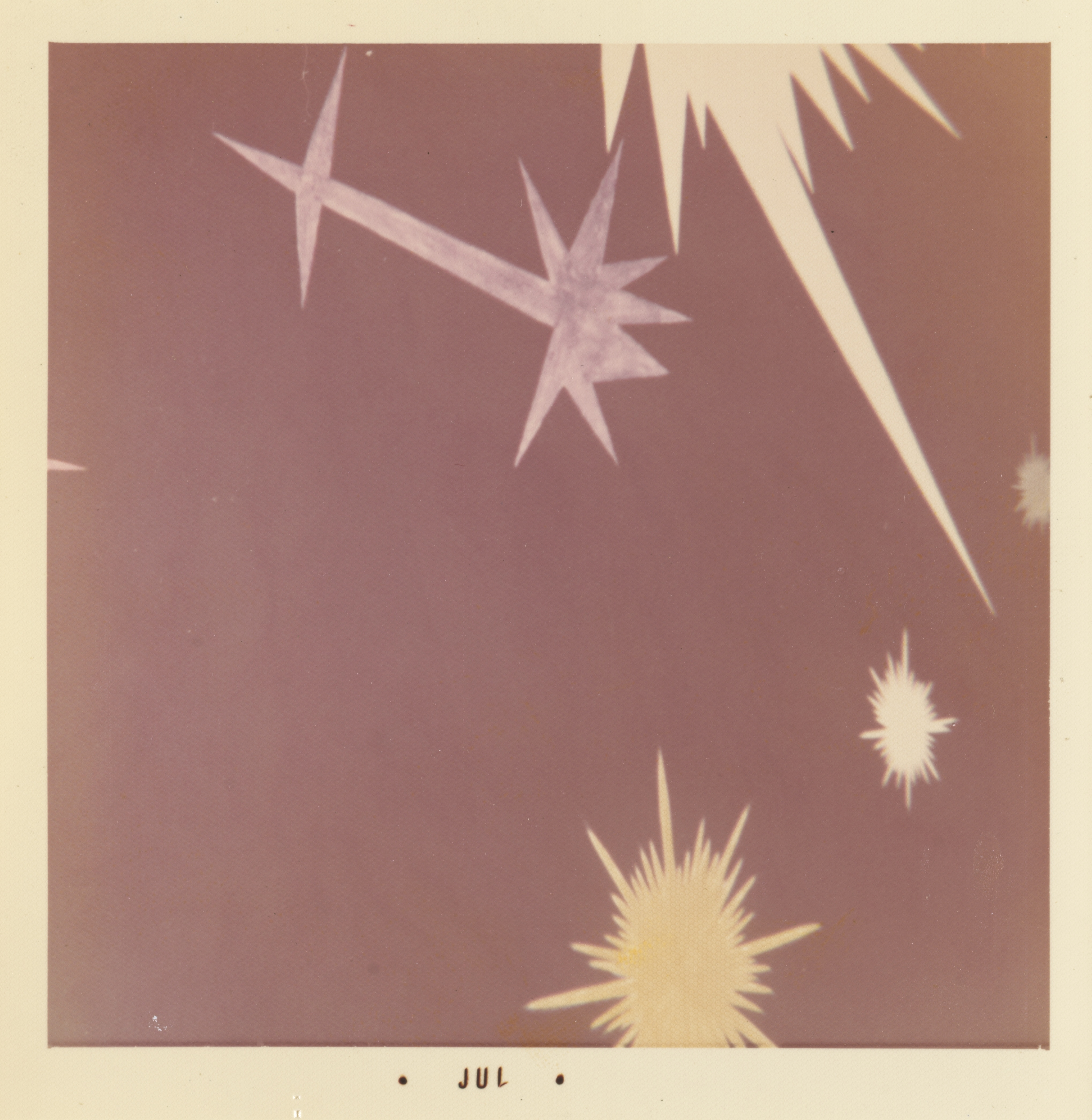 Photograph of a stylized starburst pattern on a mauve background. The design features several sharp, radiating shapes in shades of white and pale yellow, including a large eight-pointed star with a long tail at upper left and jagged bursts at the top and bottom right. The white border of the print includes the text “• JUL •” centered along the bottom edge.