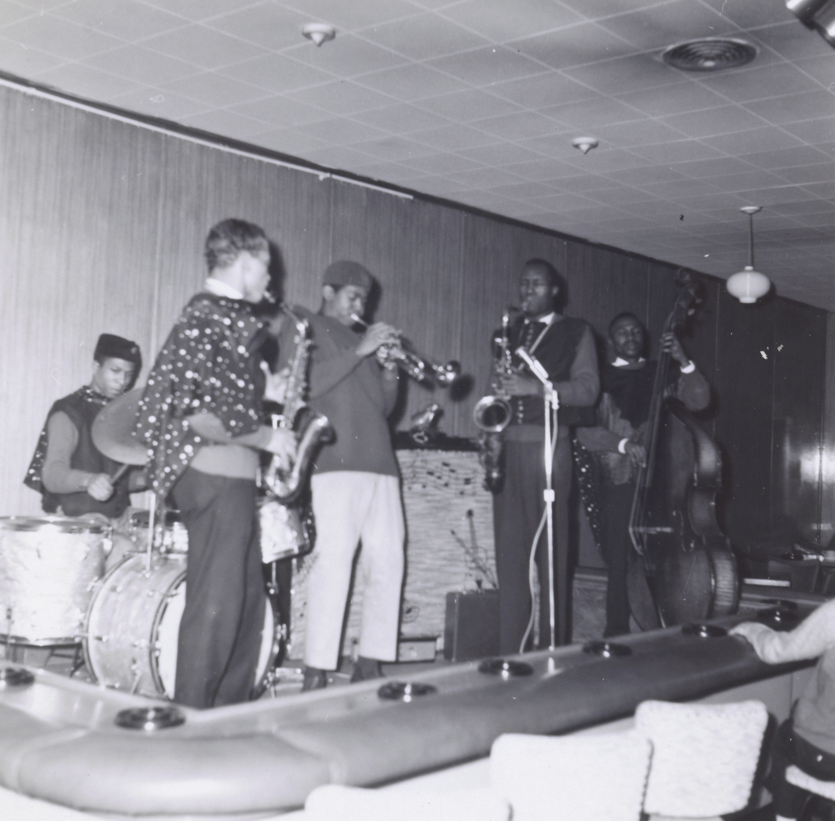 Photograph of a five-piece band performing on a stage in a lounge with upholstered seating in the foreground. From left to right: a drummer in a sleeveless shirt and fez-style hat, a saxophonist wearing a polka-dotted shirt, a trumpet player in a fez and light pants, a second saxophonist in a dark sweater, and a bassist smiling while playing an upright bass. A hanging light fixture and ceiling vent are visible above the stage area.