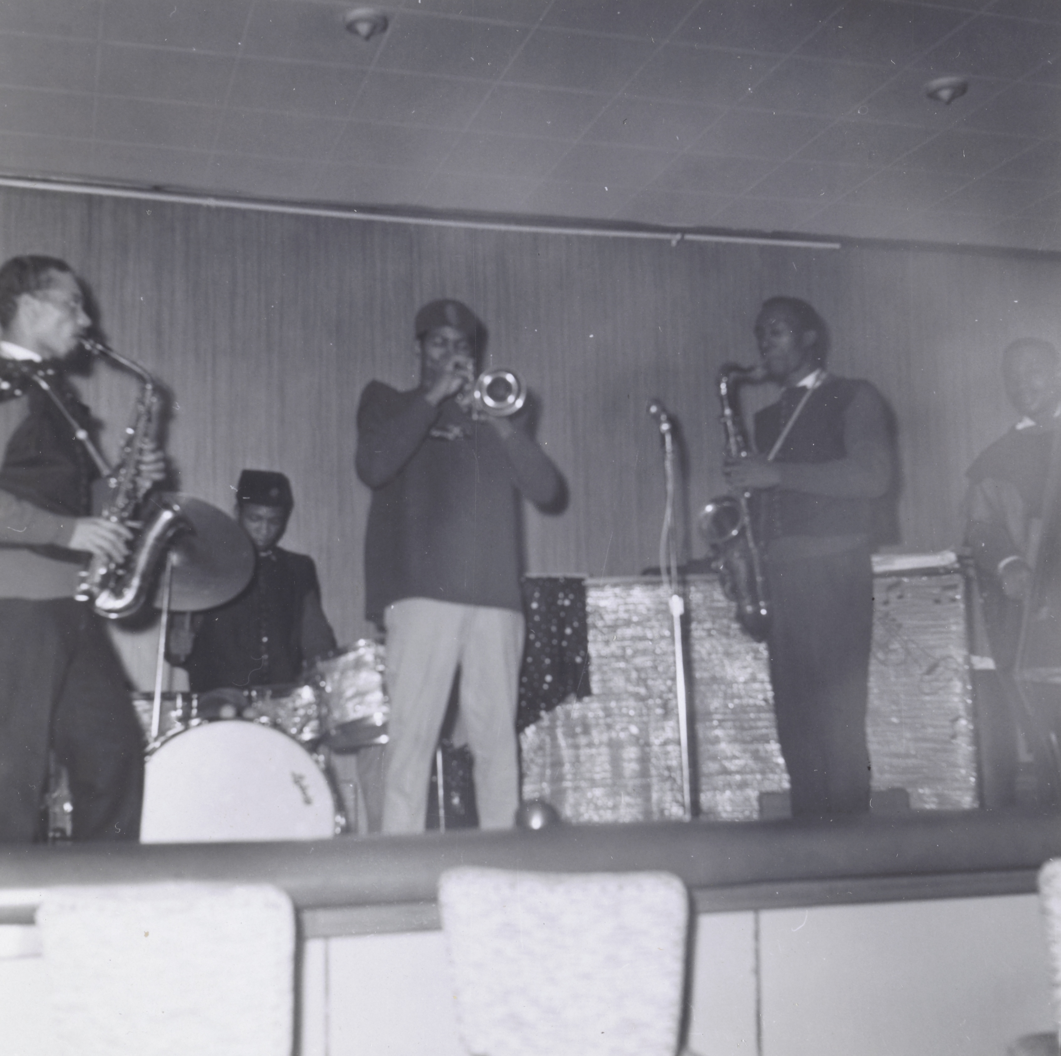 Photograph of six musicians performing on a low stage with a wood-paneled wall and a drop ceiling. From left to right: a man playing a saxophone, a drummer behind a white bass drum, a trumpet player wearing a fez-style hat and dark shirt, another saxophonist in a sleeveless sweater, and a bassist at far right playing an upright bass. Two microphones on stands are positioned at the front of the stage. Upholstered chairs are visible in the foreground.