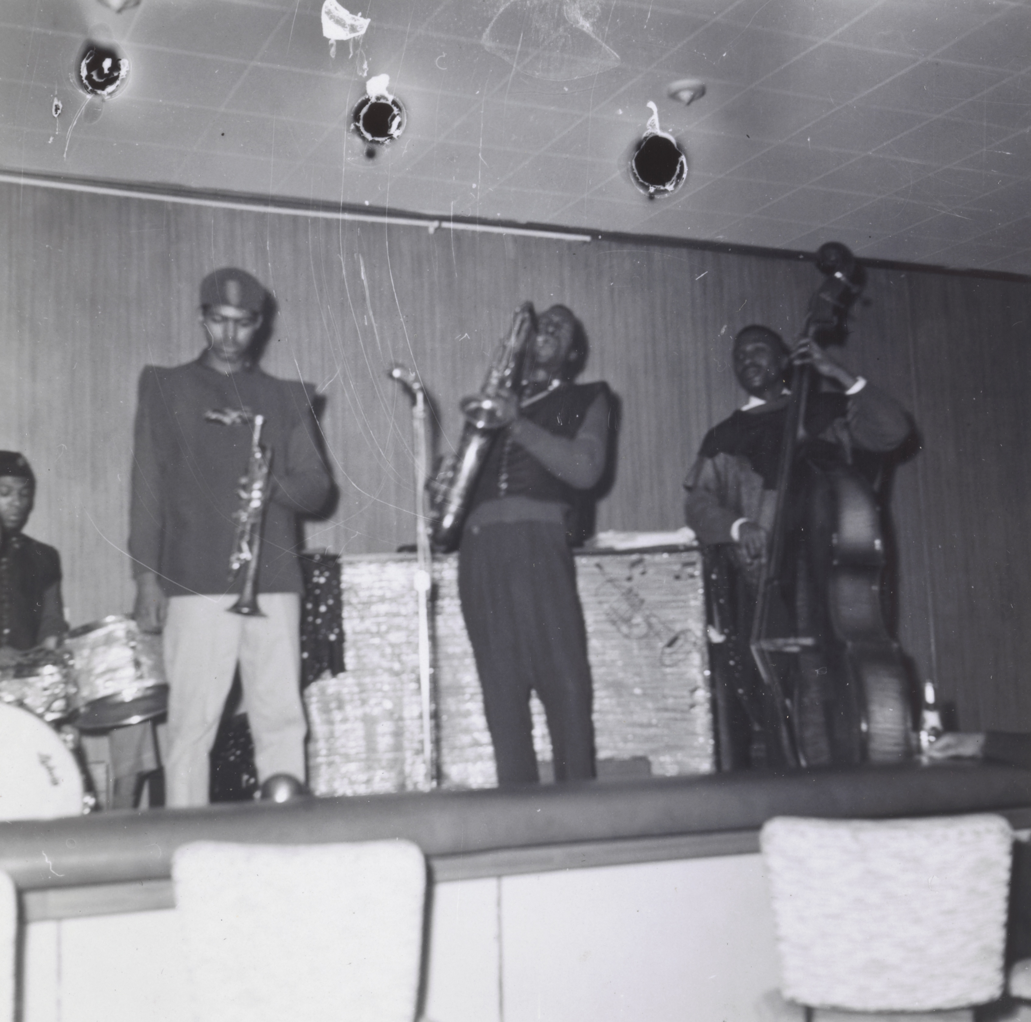 Photograph of a five-piece band performing on a low stage in front of a wood-paneled wall. From left to right: a drummer partially visible behind a white bass drum, a man in a dark shirt and fez-style hat holding a trumpet at his side, a saxophonist playing mid-note with his head tilted back, a bassist holding an upright bass, and a keyboard player mostly obscured at far right. The ceiling has multiple damaged areas with torn material exposing black holes. Upholstered chairs are visible in the foreground.