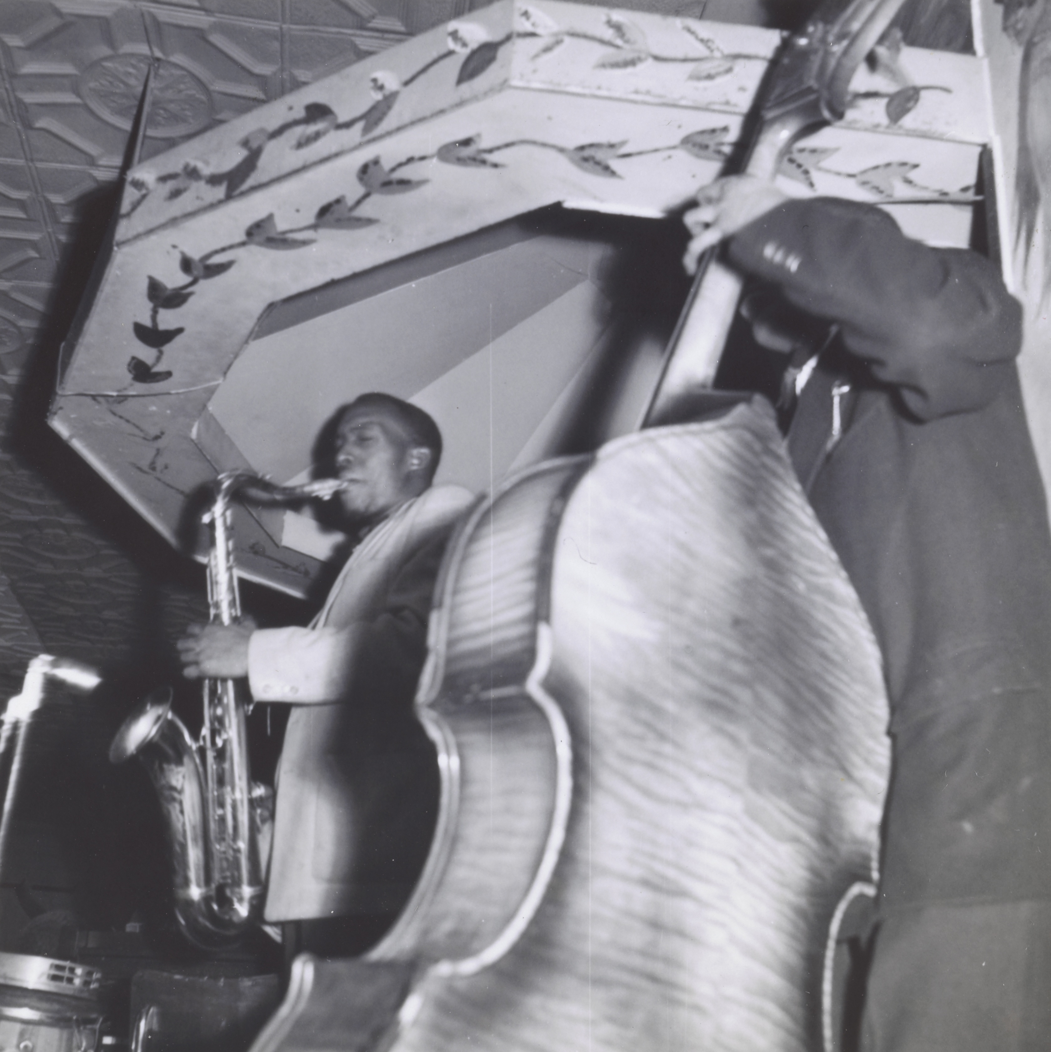 Photograph of two musicians performing on stage, viewed from a low angle. At left, a man in a light jacket plays a tenor saxophone beneath a canopy decorated with a leafy vine pattern. At right, a second man plays an upright bass, the large curved back of the instrument filling much of the frame. An ornate, coffered ceiling is partially visible above them.