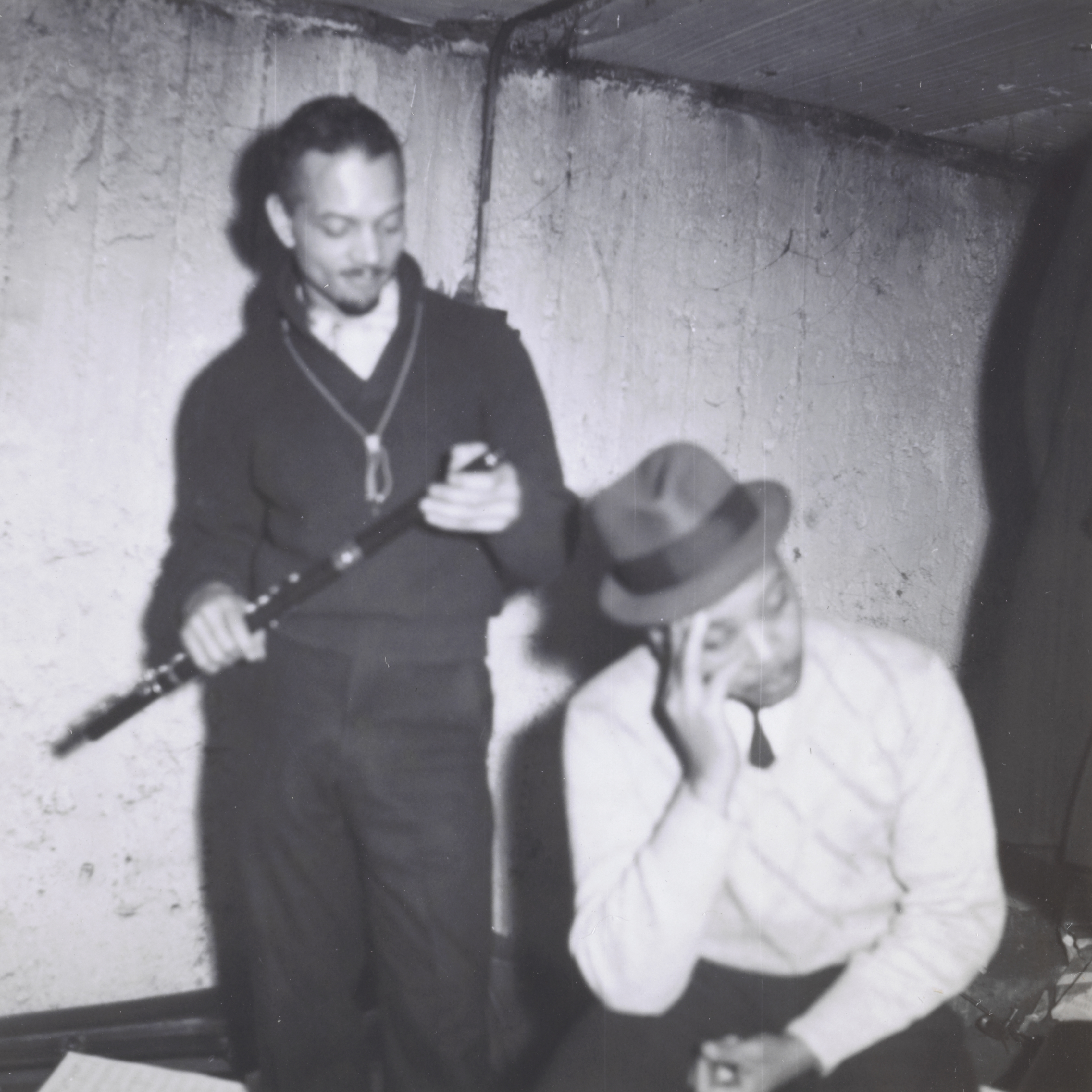 Photograph of two men in a bare room with textured concrete walls. At left, a man stands holding a flute, looking down at it with a slight smile. He wears a dark sweater and slacks, with a light shirt collar visible at the neck. At right, another man in a light sweater and fedora sits on a bench, resting his head in one hand and looking downward. A page of sheet music lies on the floor in the lower left corner.