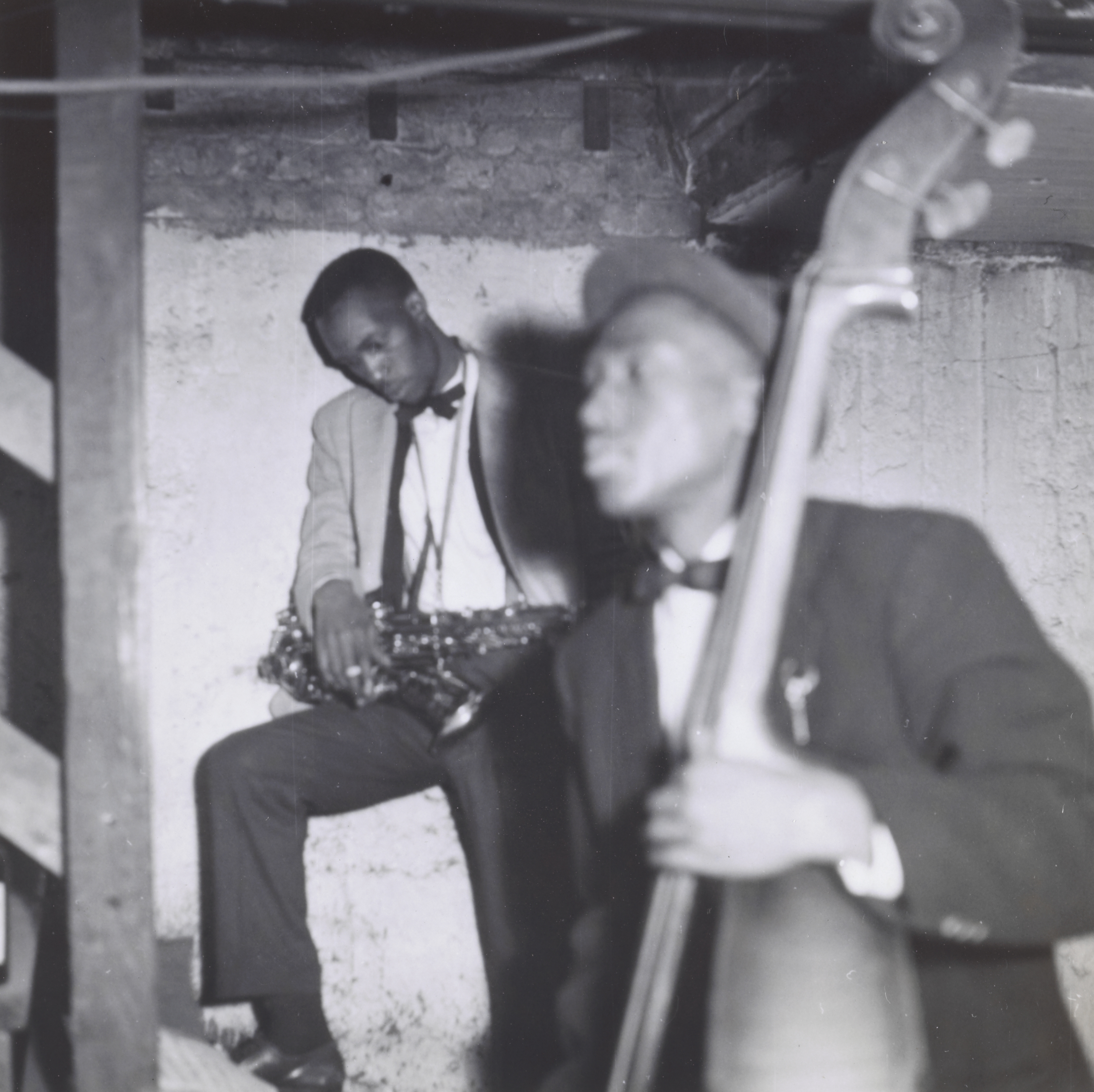 Photograph of two musicians in a rough, unfinished basement space. At left, a man in a light jacket and bow tie sits on a ledge, resting a saxophone across his lap and looking down. In the foreground at right, another man in a dark suit and bow tie plays an upright bass, his face blurred in motion. Exposed brick and wooden beams are visible behind them.