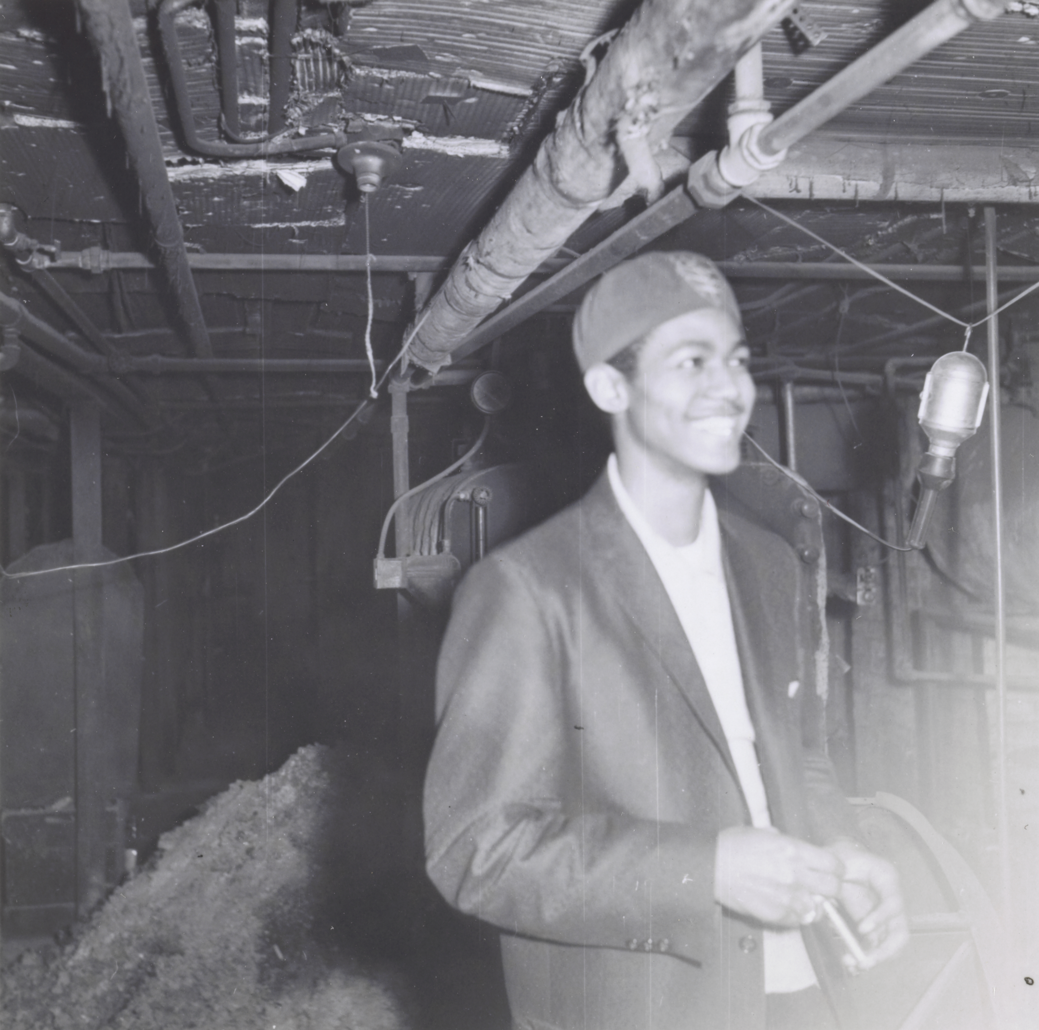 Photograph of a young man standing in a dimly lit basement with exposed pipes overhead. He wears a dark jacket, light shirt, and a fez-style hat with an emblem. He smiles while looking slightly off camera, holding an object in his hands. Behind him, industrial equipment and a pile of loose material are partially visible in the background.