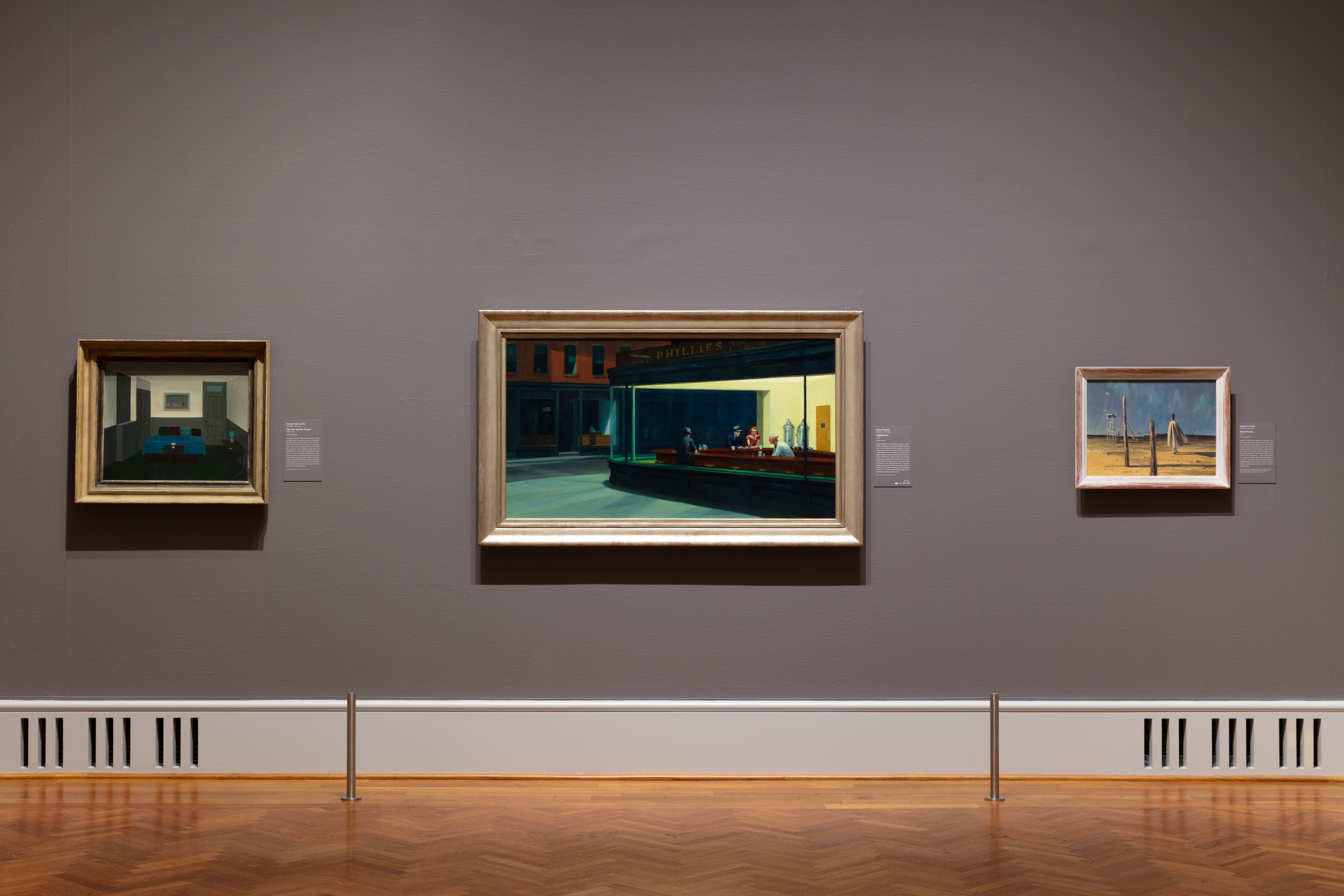 On a gray gallery wall, from left to right, are Gertrude Abercrombie's "The Past and the Present": a painting of a small, sparse room with a daybed;  Edward Hopper's "Nighthawks": a painting of four people in an urban diner at night; and Hughie Lee-Smith’s "Desert Forms": a painting of a desert through which a figure walks with their back to the viewer, cape billowing.