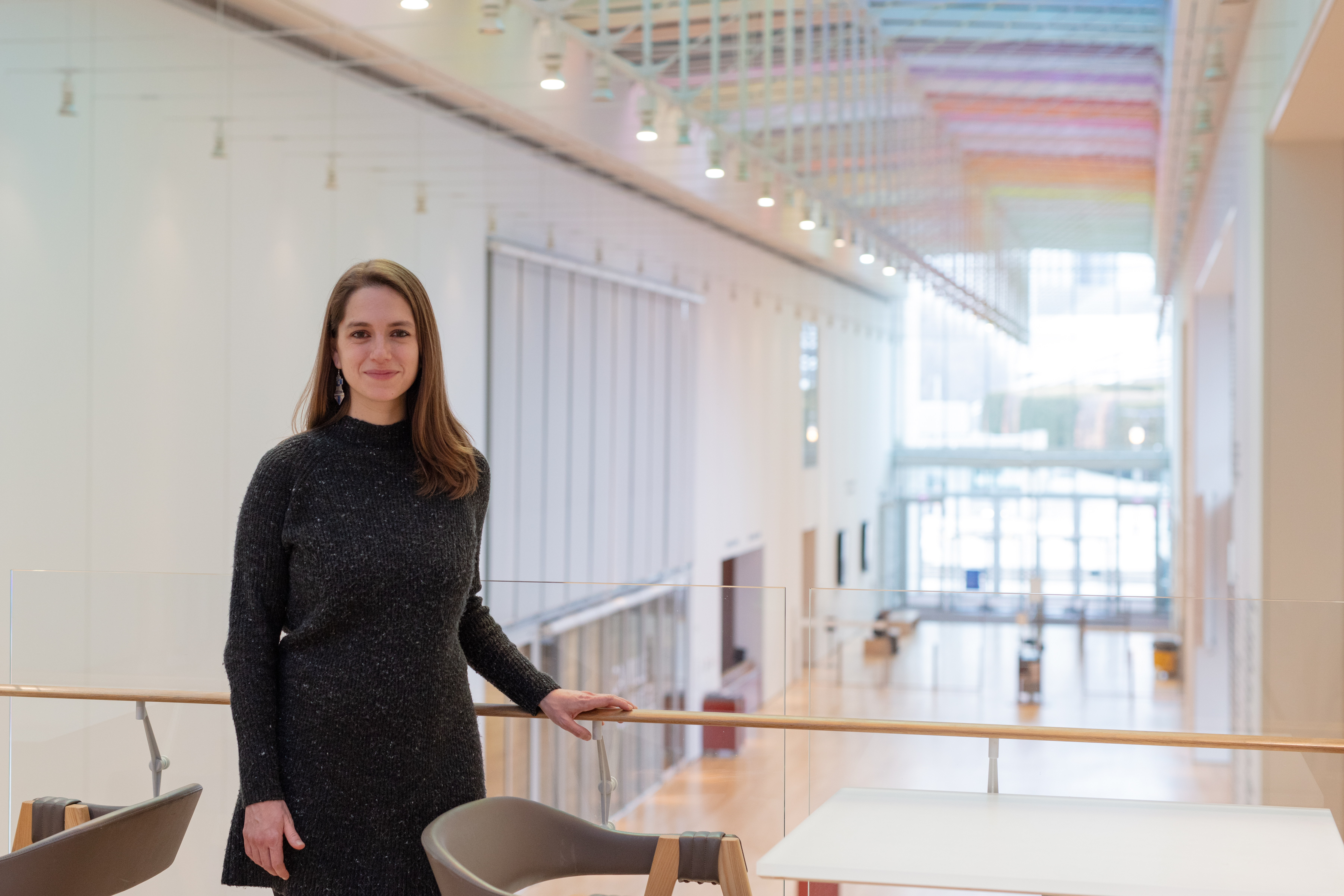 A photo shows a young, light-skinned woman with light-brown hair smiling and facing the viewer. Behind her and to her right is a balcony rail, beyond which extends the light-filled, white expanse of the Art Institute of Chicago's Griffin Court, within the Modern Wing. Glass doors and windows hint at a park just beyond.