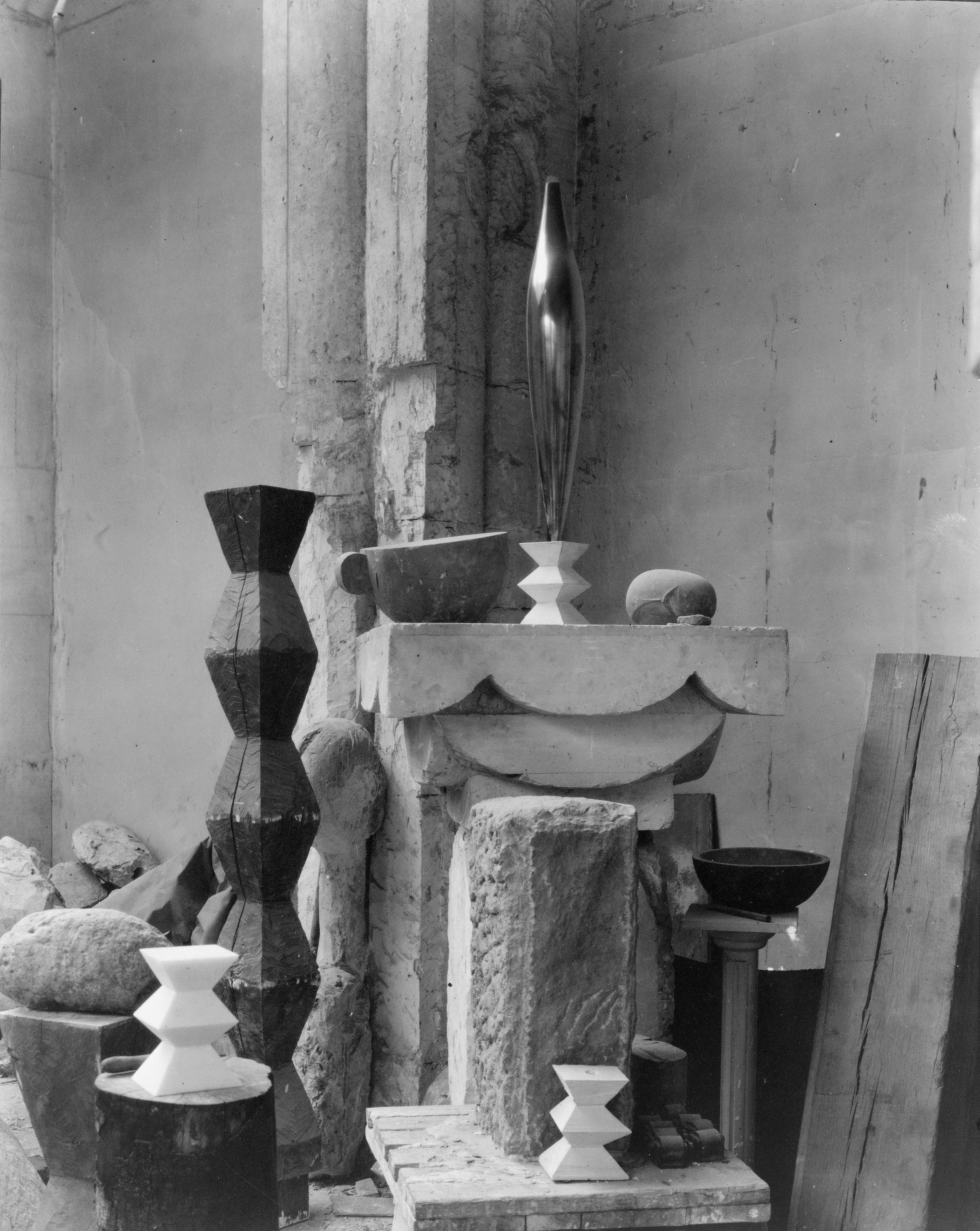 Black-and-white photograph of a room corner with arranged sculptures: at center a tall shiny vase-like form on a white hourglass base atop a ledge; at left a dark faceted column and stones; below and front rough stone blocks and small white hourglass forms; at right a bowl and leaning boards.