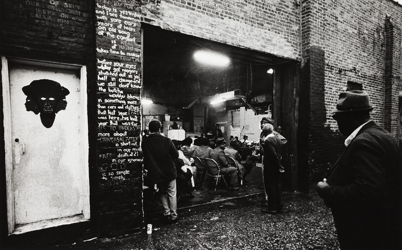 Black and white photograph looking from an alley into the Garage, where people are sitting and listiening to someone perform