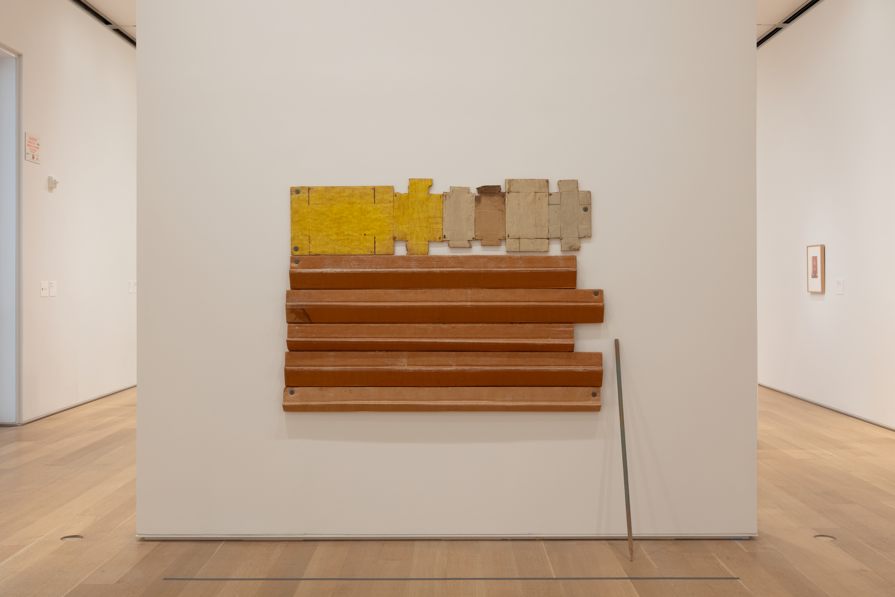 Photo shows a generally rectangular sculpture affixed to a white gallery wall. A horizontally corrugated bottom section appears almost coppery. The upper portion, yellow at left and shades of beige at right, could almost be a string of opened-up boxes.