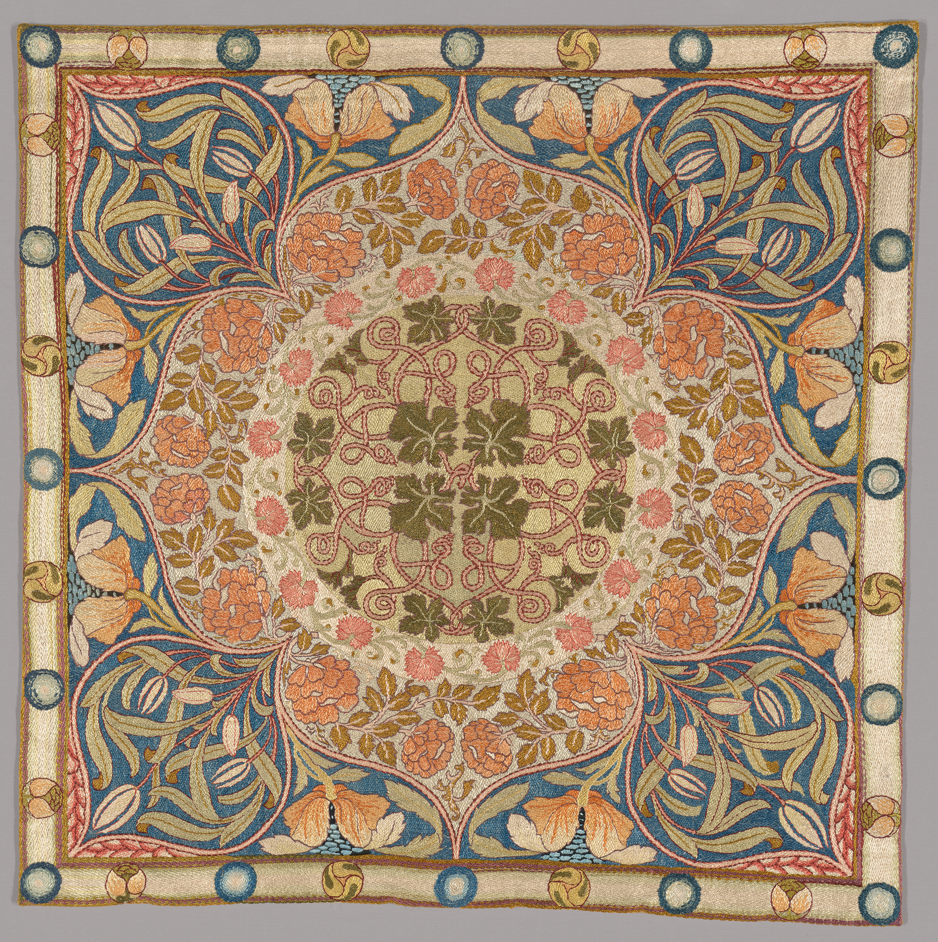A square tapestry in beige, pink, peach, green, and blue featuring a central, four-leafed blossom. Inside the blossom is an intricate floral motif with leaves and geometrically twisted vines at center.