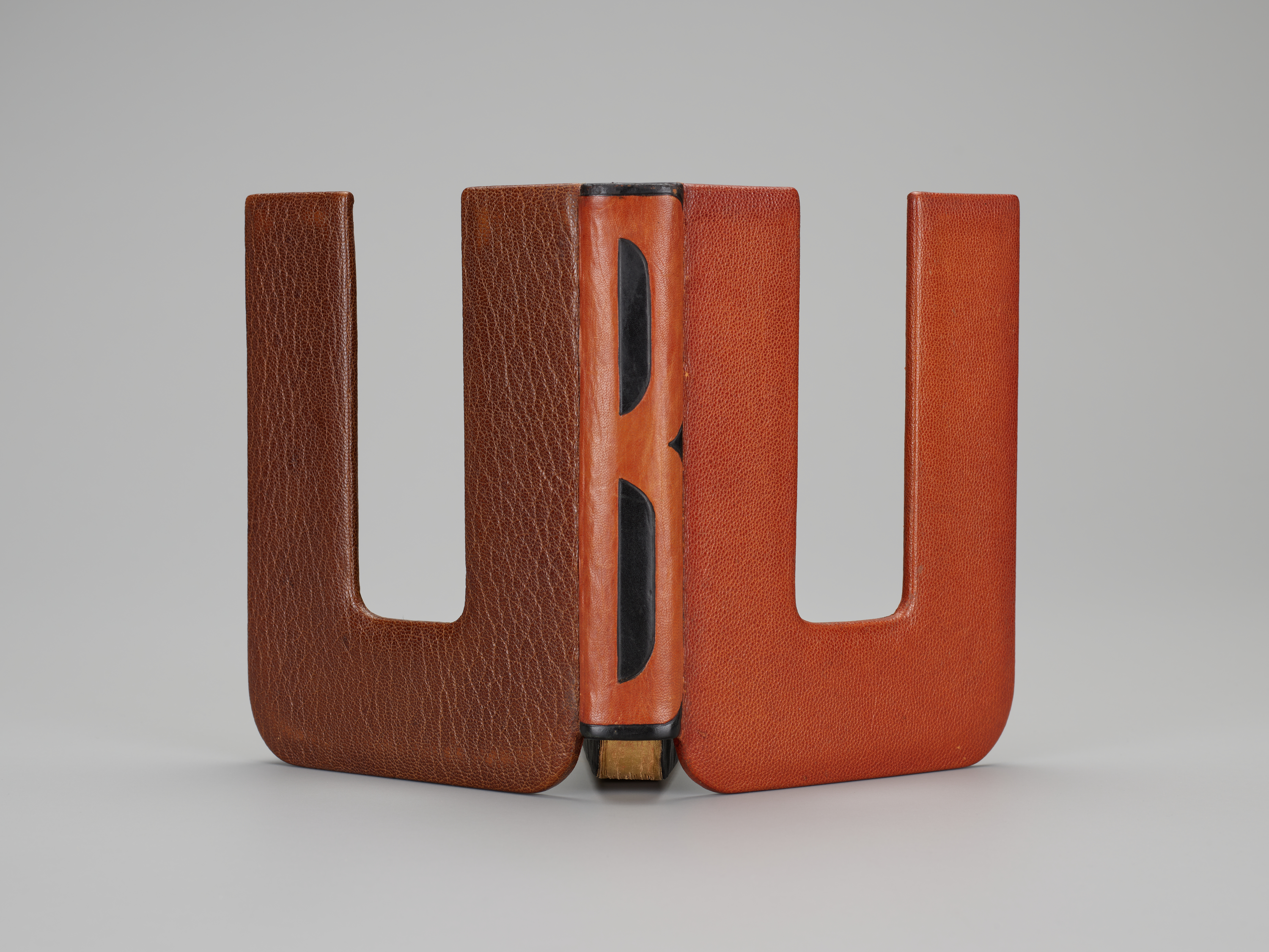 A leather-bound book features a cover cut into a "U" shape, both front and back. The book is open and standing so that the viewer sees both covers and the spine, but not the pages. The spine is decorated in black in in such a way that the exposed leather suggests the letter "B". Together, the covers and spine seem to read "UBU."