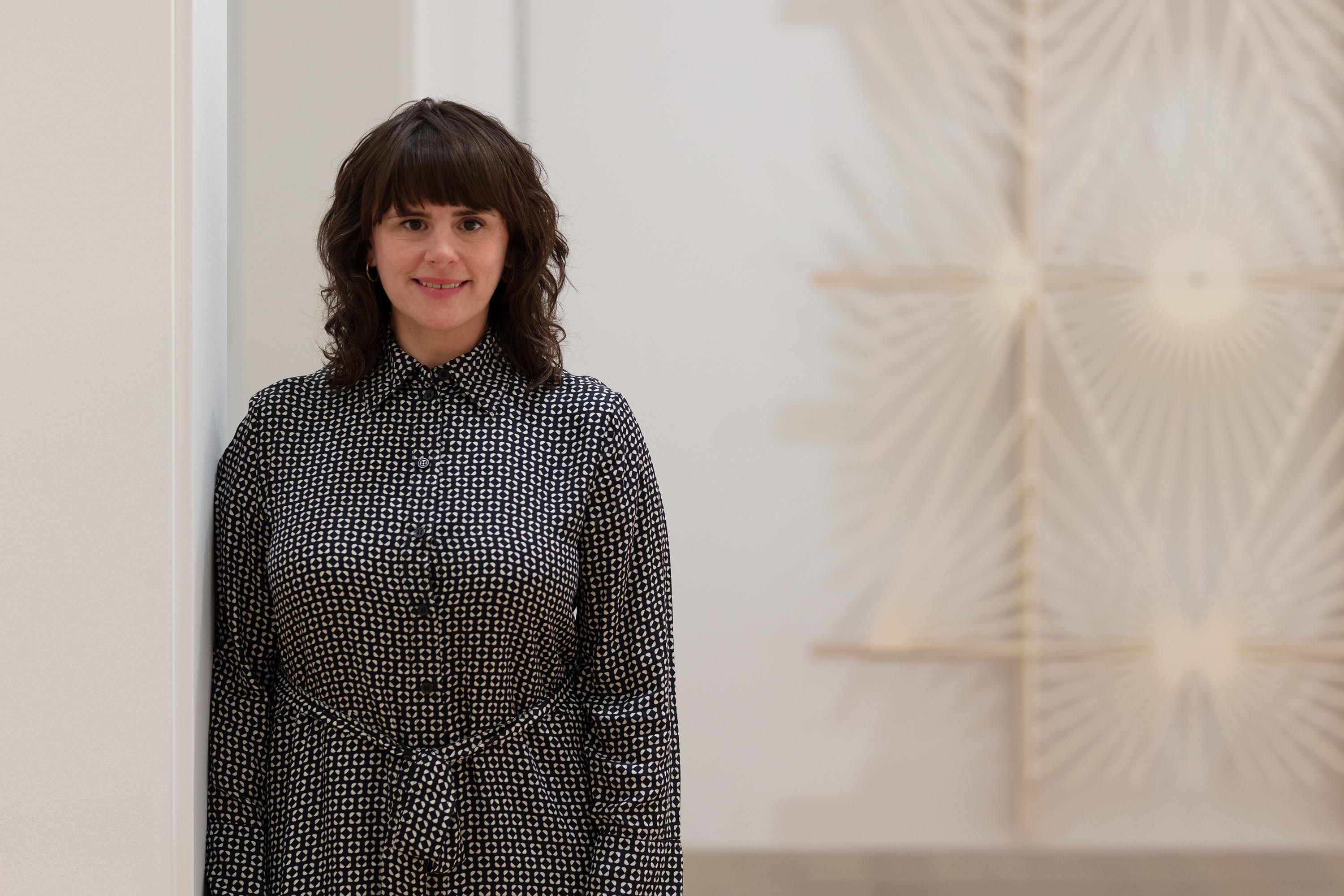 Photo of Alison Fisher, a light-skinned woman with shoulder-length, light-brown hair and bangs. She smiles, looks to the viewer, and leans slightly against a white gallery wall. She wears a black and white patterned shirtdress.
