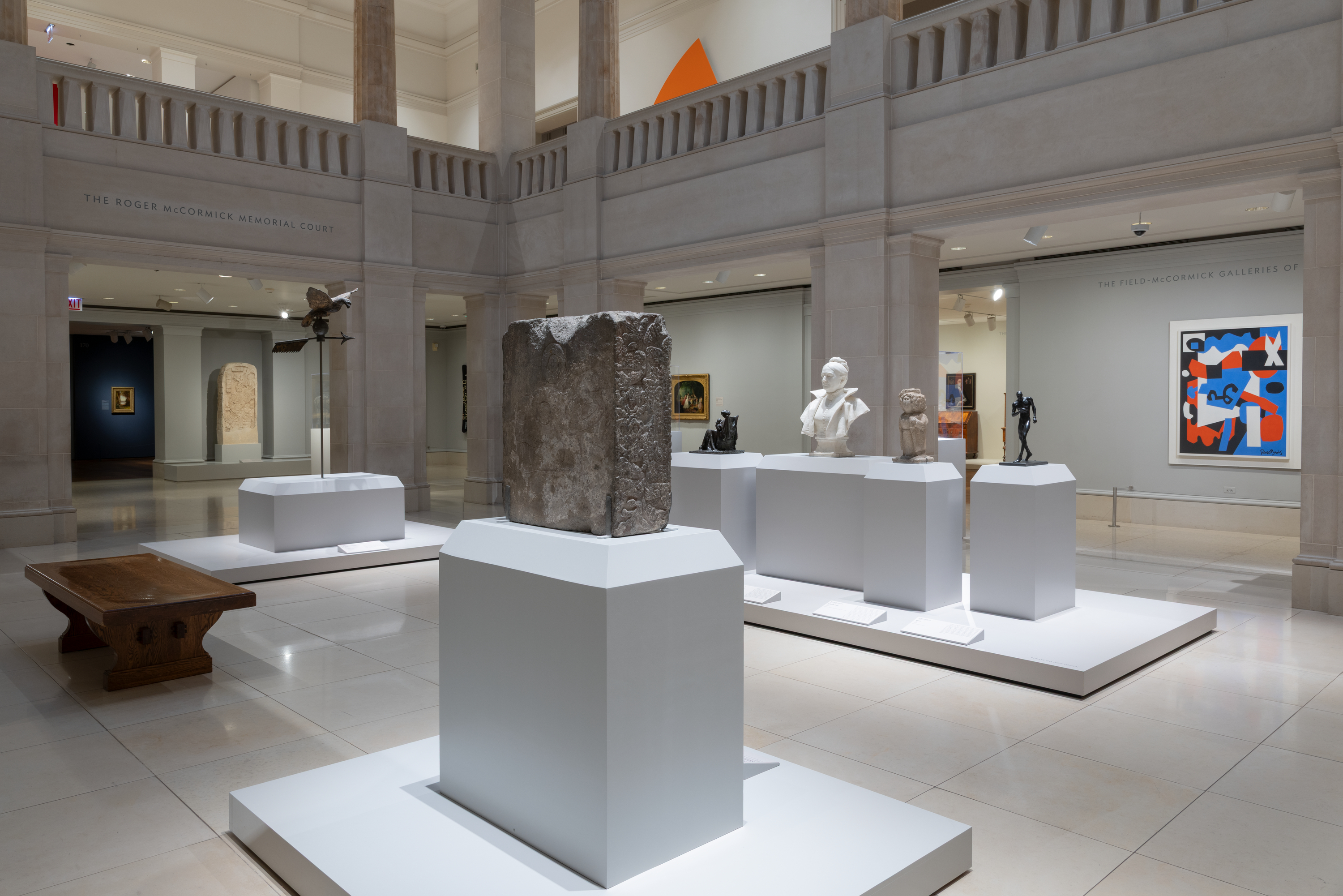 Color photograph of a museum gallery shows a rough gray stone block on a white pedestal at center, with white plinths holding busts and small sculptures behind it, a low wooden bench at left, beige stone columns around, and a blue, orange, and white abstract painting at right.