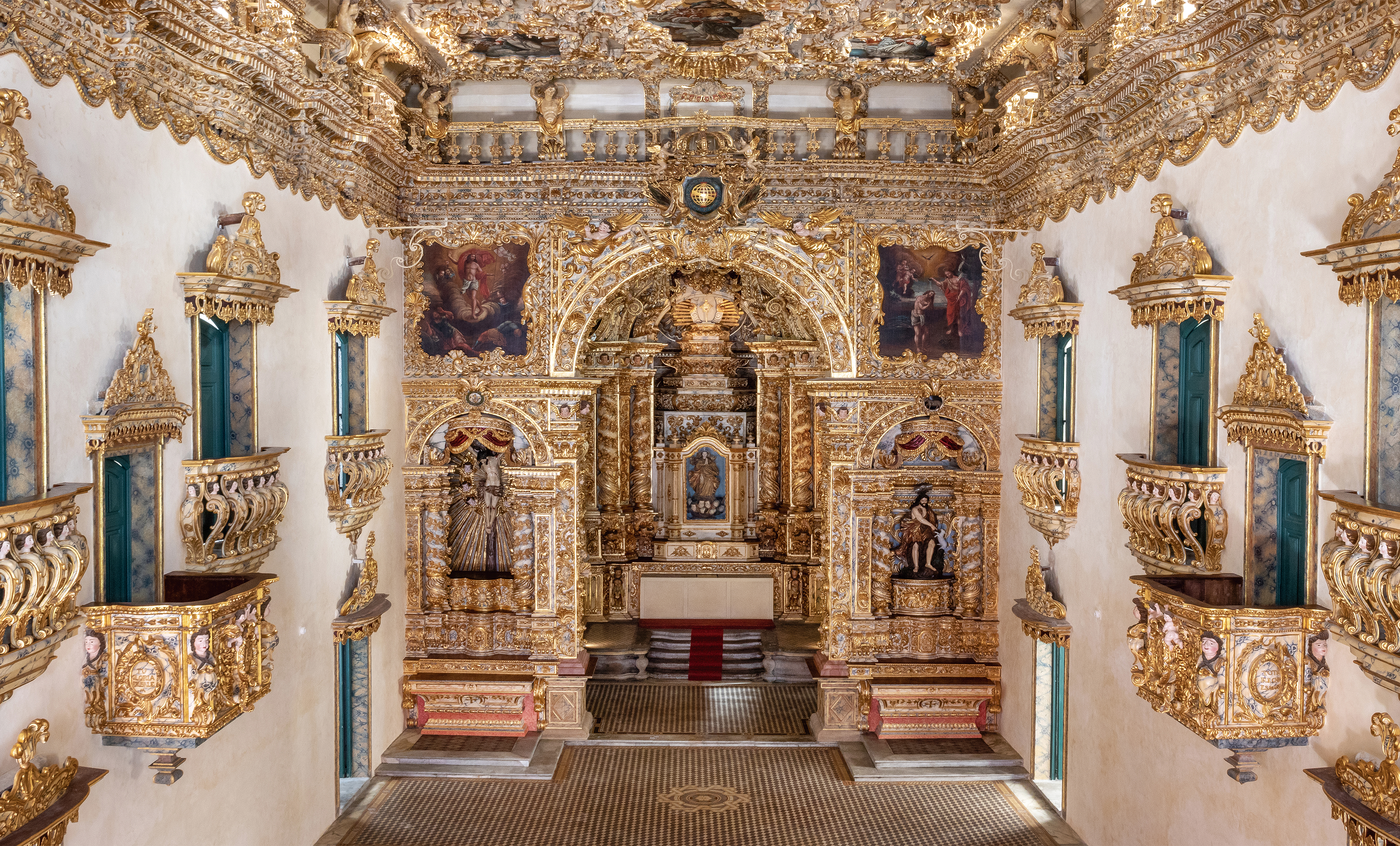 An interior of a church with an elaborate altar made of several intricately carved sculptural motifs. Two sculptures and two paintings appear on each side of the altar. Along the walls on each side are several balconies with gold railings and pediments and in front of cool green doors.