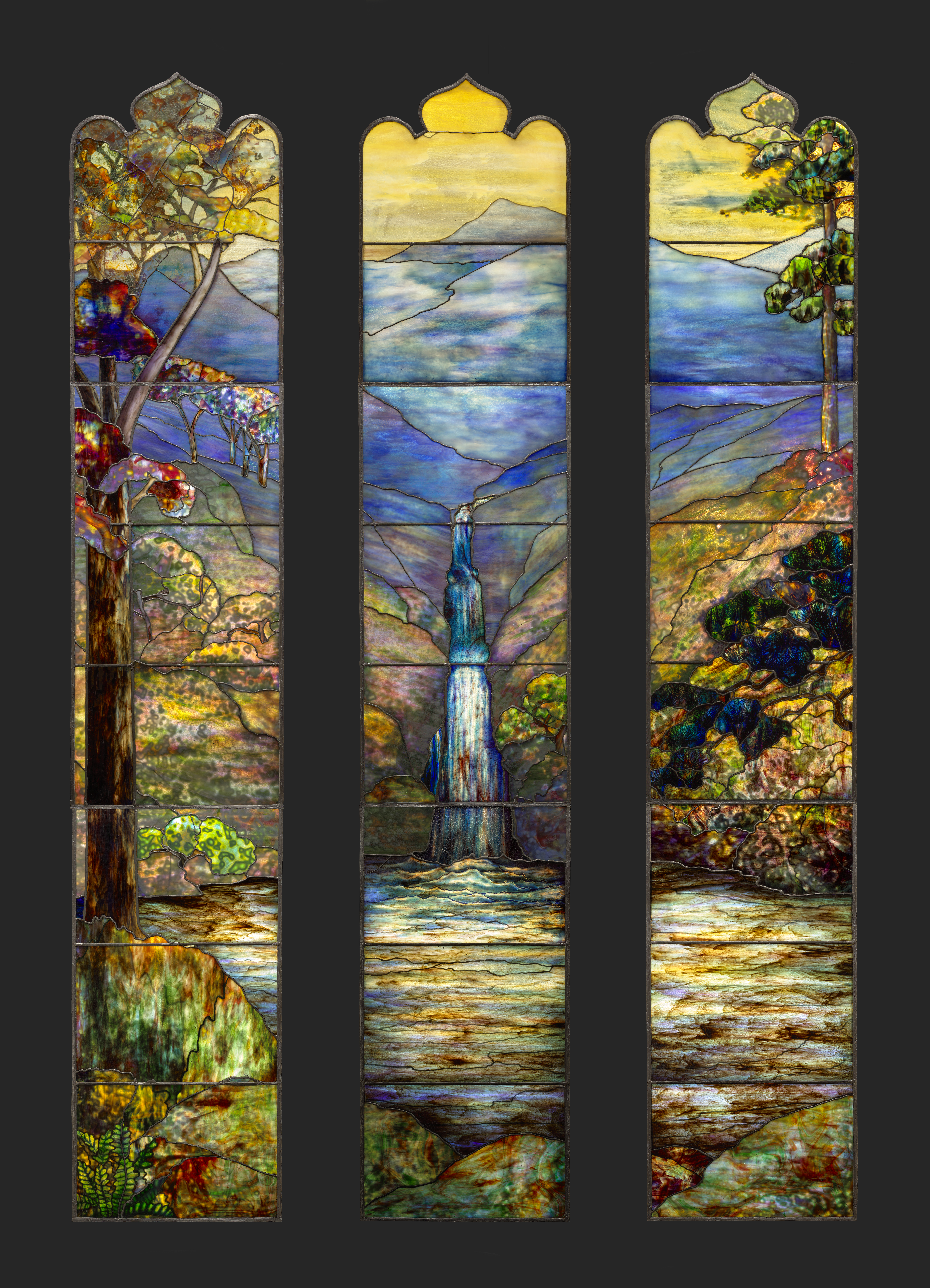 A panel of a stained glass window shows a blue and purple mountain beneath a bright yellow sky. The richly colored, decoratively shaped panel appears against a black background.