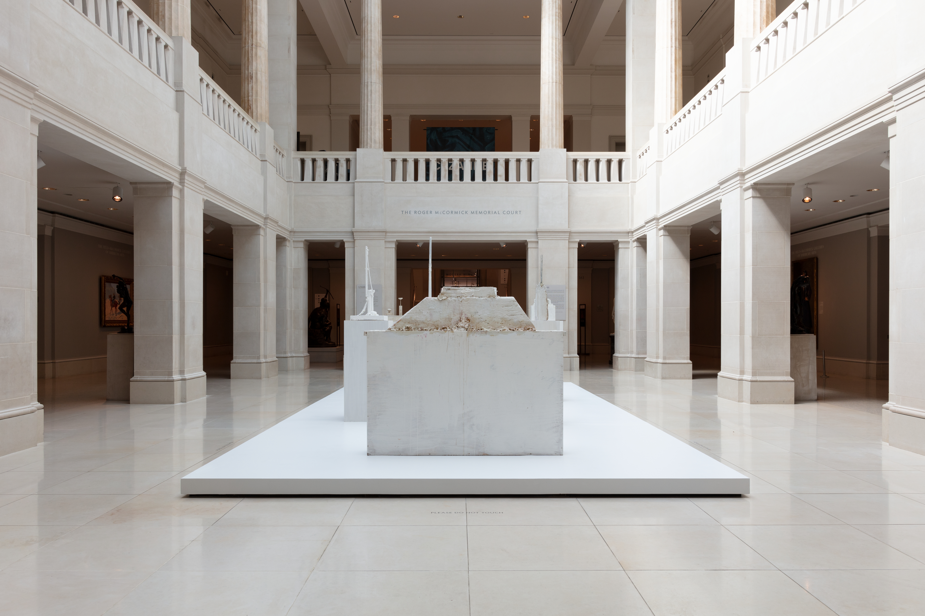 Photo of an art gallery shows a sculpture resembling a pyramid with a shorn top, made of wood and painted white. On top of the structure is a similarly shaped smaller structure.
