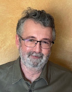 A middle-aged man with glasses and a gray beard smiles warmly. He wears a green shirt against a textured golden background, conveying friendliness.