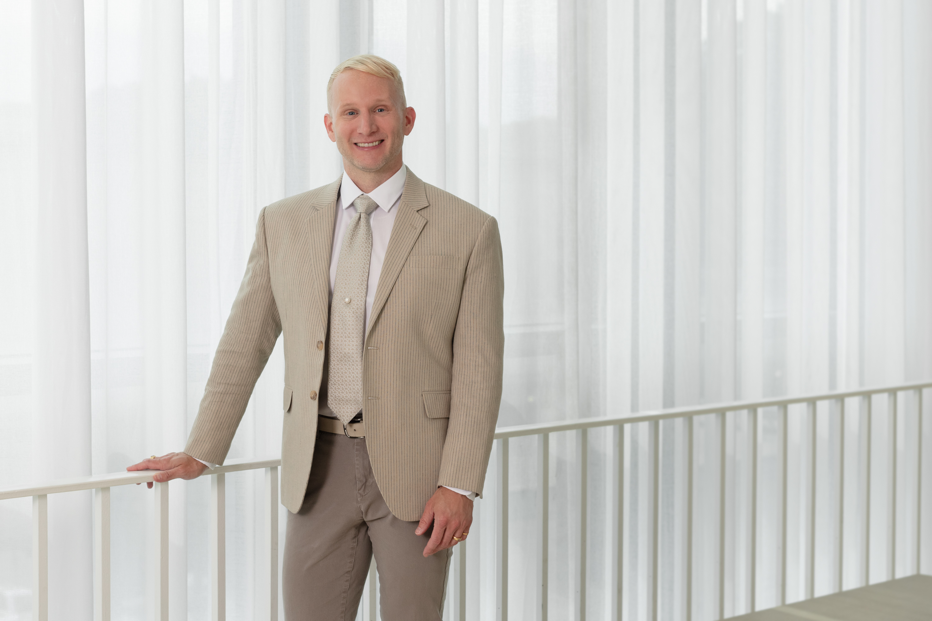Photo of curator Andrew Hamilton standing at a railing before a white curtain, smiling broadly. He has fair skin and blond hair and wears a beige pinstripe blazer, taupe pants, and a beige tie.