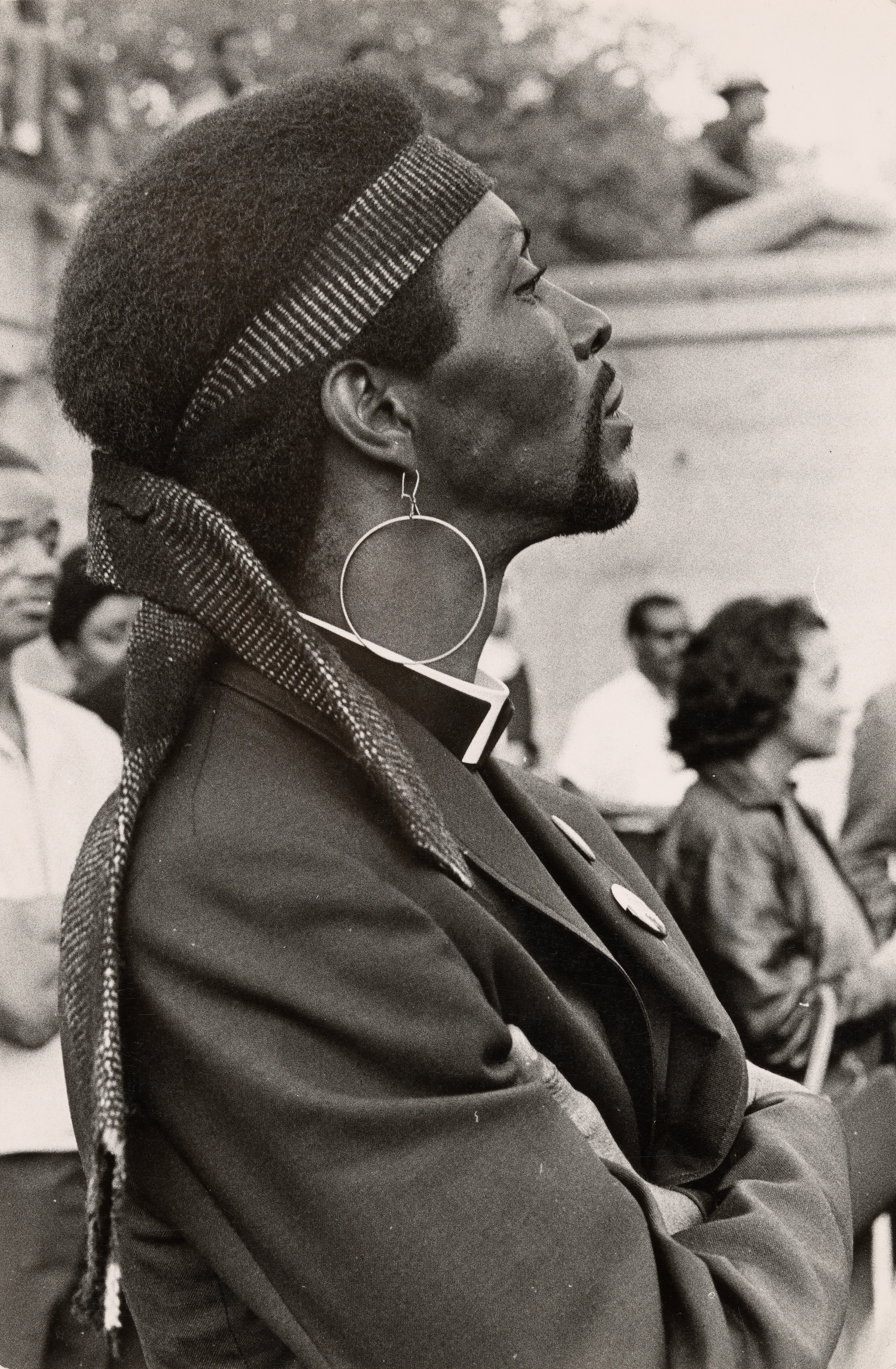 Black-and-white photograph of a man in profile standing with arms crossed, facing left. He wears a clerical collar, a dark jacket with pinback buttons, and a wide striped headwrap tied at the back. A large hoop earring hangs from his left ear. His hair is styled in a rounded Afro, and he has a neatly groomed beard. Several people are partially visible in the blurred background, suggesting a public gathering or event.
