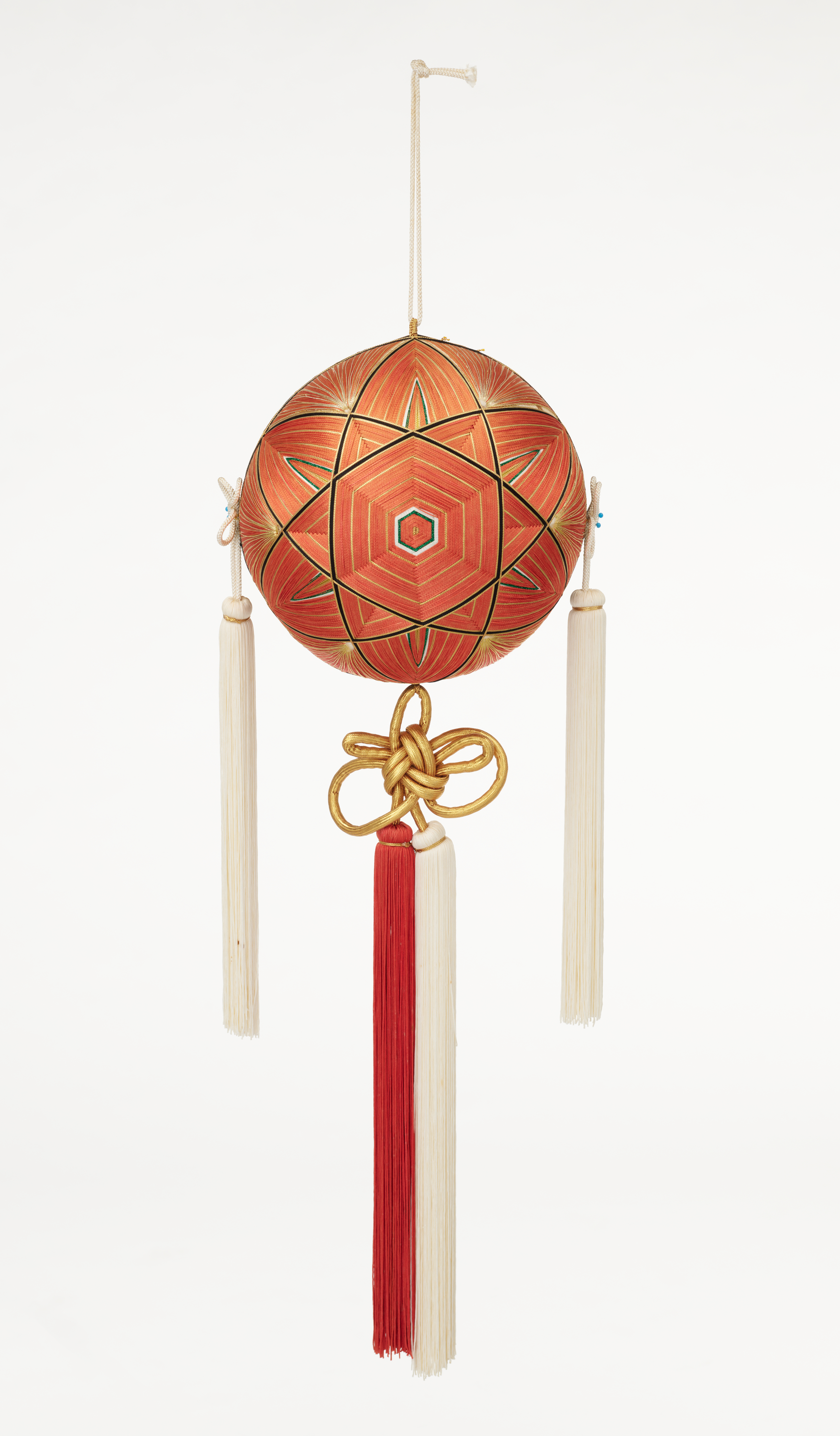 An ornament comprises a ball with a graphic hexagon/star-shaped design in shades of orange and tassels in white and orange that hang on each side and from the bottom.