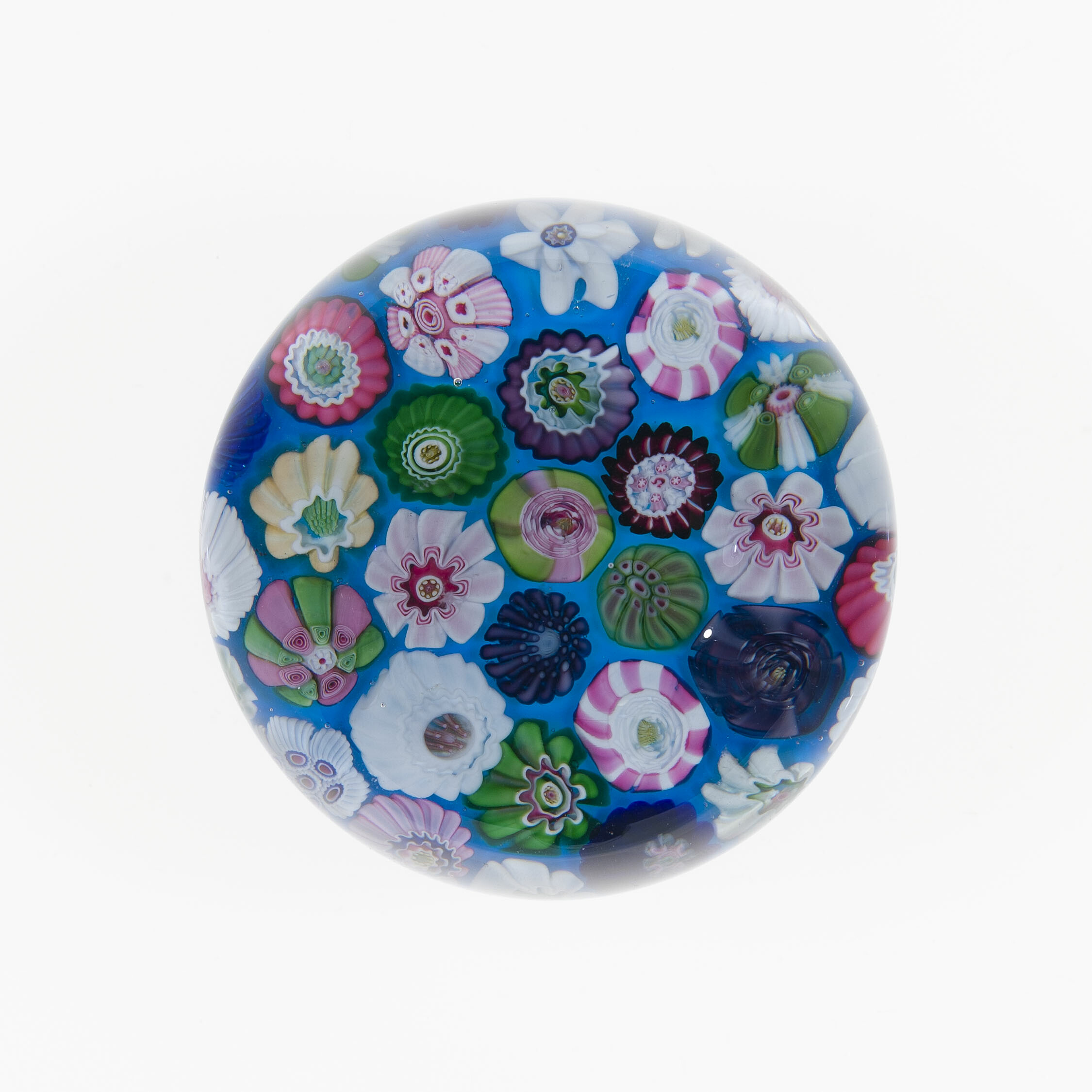 A color photograph shows a round glass paperweight at center on a white background, its blue interior filled with small flower shapes in pink, green, purple, white, and yellow from left to right, with several white blooms at top and a dark blue swirl at lower right.