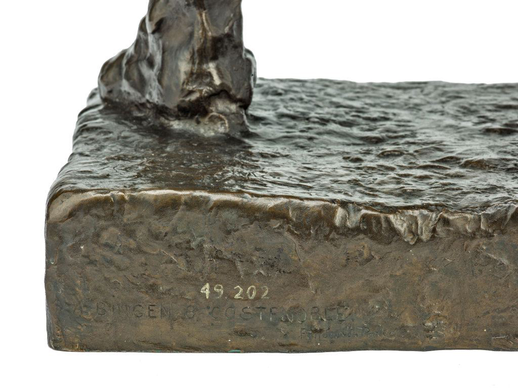 Foreground shows a dark brown rectangular base with a rough, rippled surface and a slightly uneven top edge; the front face has pale numbers “49 202” and faint, partially legible letters along the lower edge. At the top left, a lumpy protrusion rises from the base, while the right side is a broad flat area extending to the edge. Background is plain white.