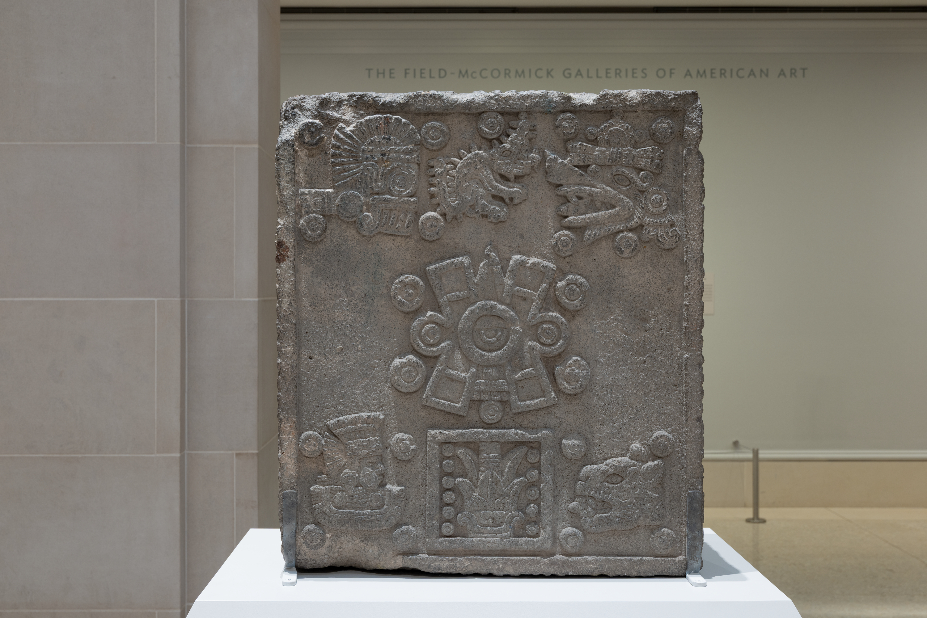 A color photograph shows a gray carved stone slab on a white pedestal in a gallery; at center is a circular emblem with radiating panels and small round dots, at top left and top right are profile heads with open jaws, and at bottom are rectangular motifs and another head at right.