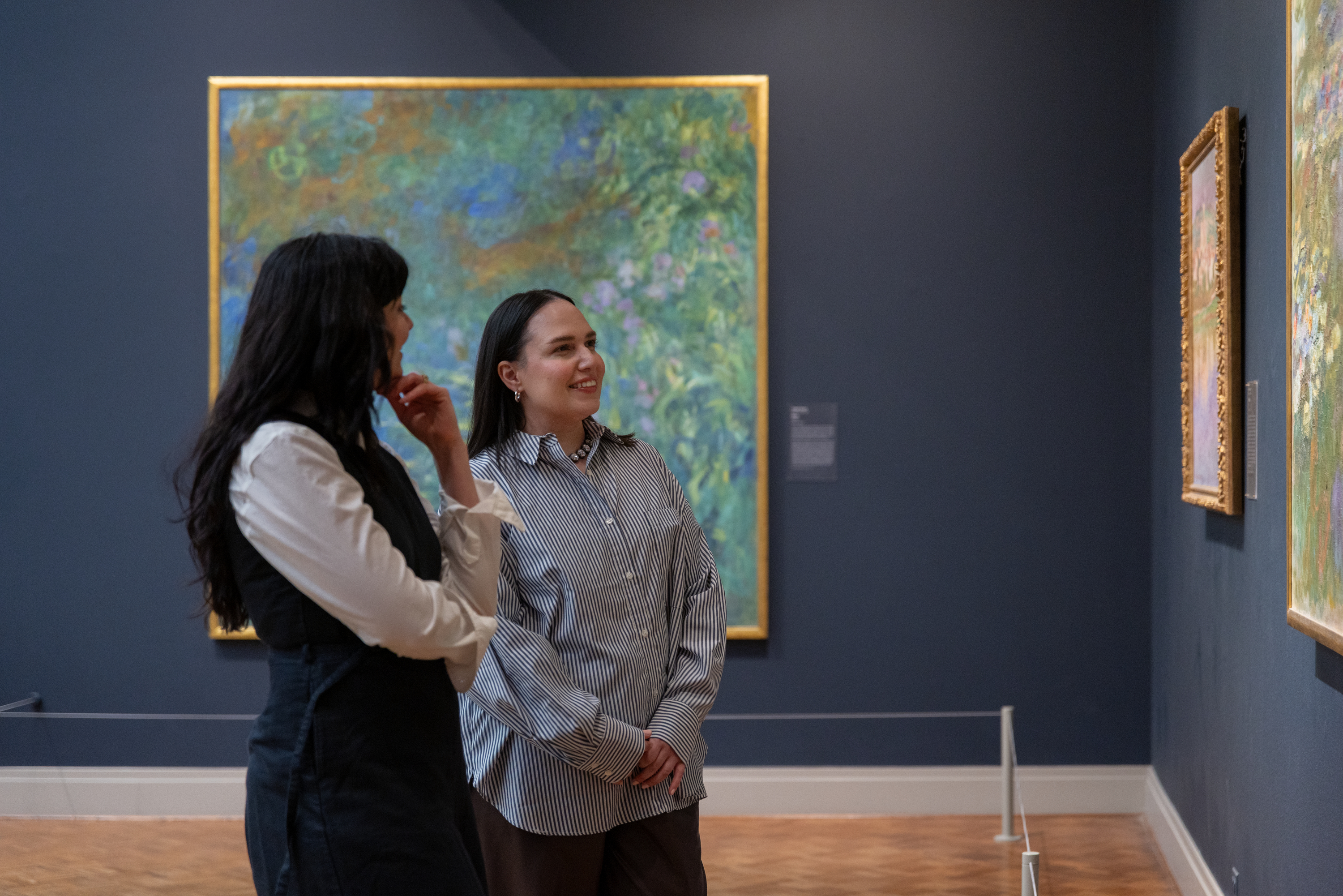 A color photograph shows a gallery with a large blue-green painting in a gold frame at center; at left and center-left stand two women, Sam Grassi and Zahra Bahia; Sam has long dark hair and wears a black dress over a white shirt, while Zahra wears a gray striped shirt. Smaller gold-framed paintings hang at right on dark-blue walls.