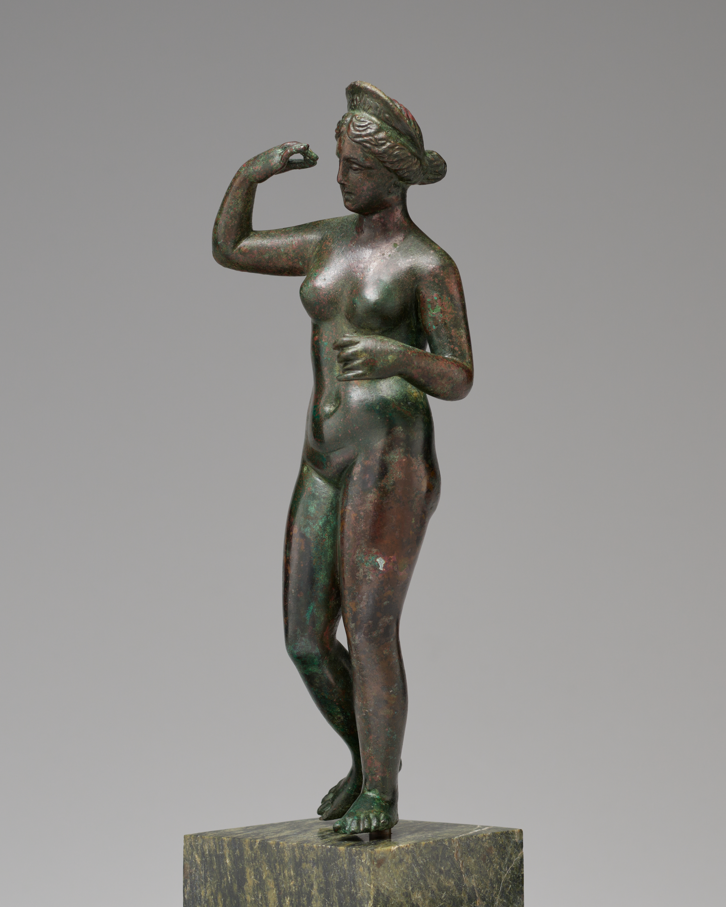 Profile view of a bronze sculpture with green patina of a nude woman with her right hand raised to eye level. She wears a tiara, her hair in a bun.
