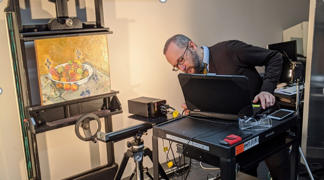 On the left, a medium-sized painting on a tall black stand shows a white bowl of red and orange fruit on a tabletop against a yellow background. In front of it, a black equipment rig with rails, a ring-shaped component, and a small tripod is positioned, with cables extending toward a cart. On the right, an adult with short gray hair wearing a dark sweater over a light blue shirt and striped tie leans over a black rolling cart holding an open black laptop, a small black box, clear glasses, a smartphone, and a red case with a yellow strap nearby.