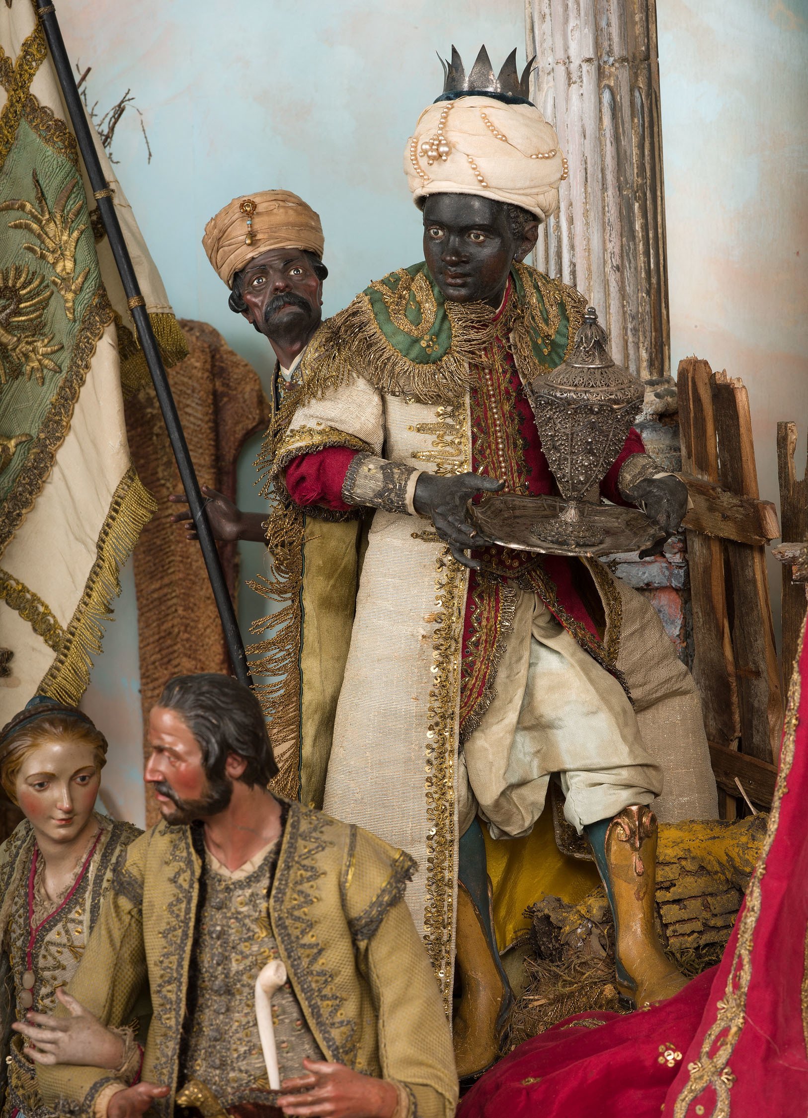 A photo of a detail of Neapolitan Creche. The photo shows multiple small figures, highlighting one of the three wisemen. The man holds an ornate object and wears a white turban and colorful