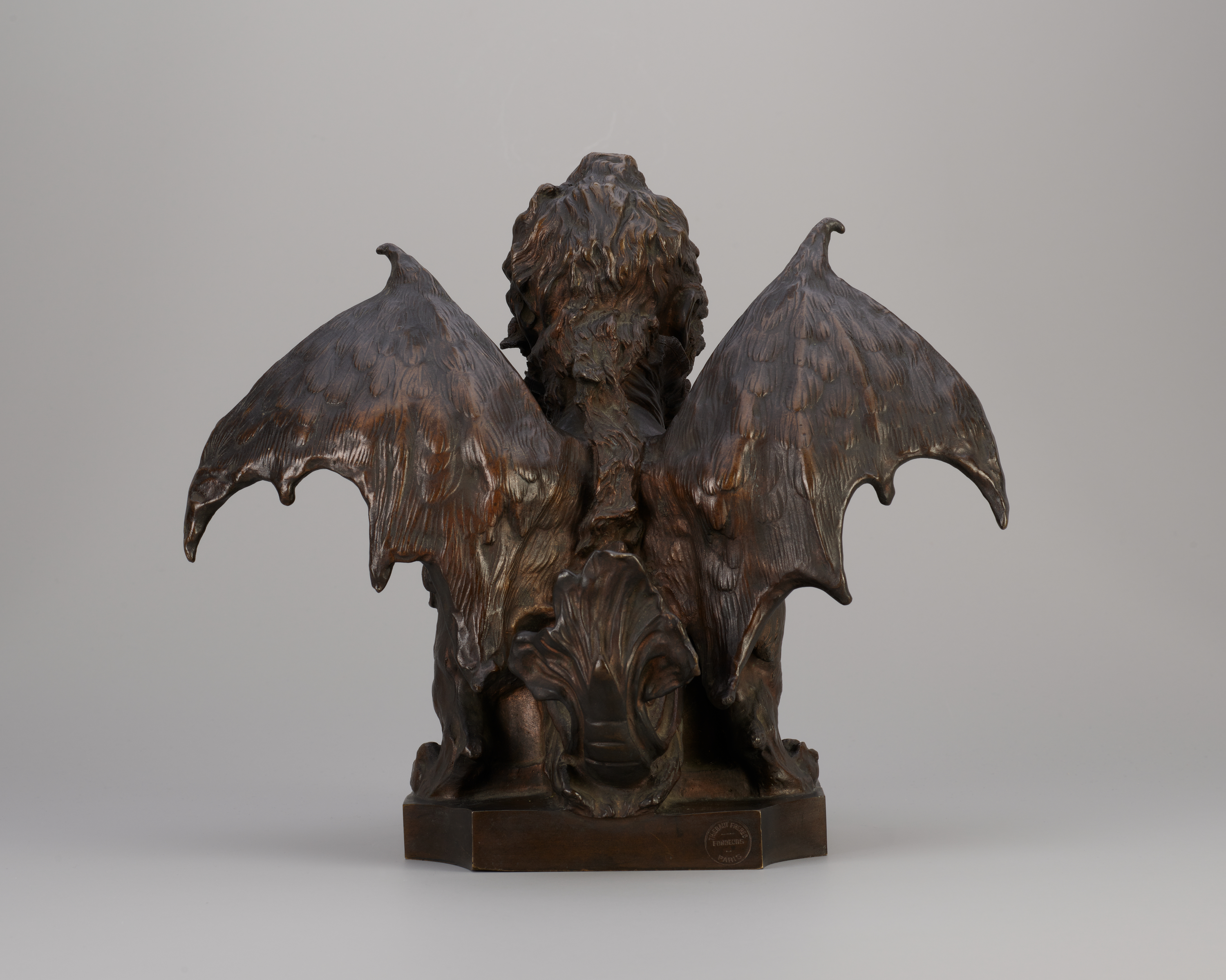 Back view of a bronze-colored metal sculpture of a creature with the head of a curly-haired person and extended bat’s wings with lightly etched feathers, a dragonlike tail curling upward at center.