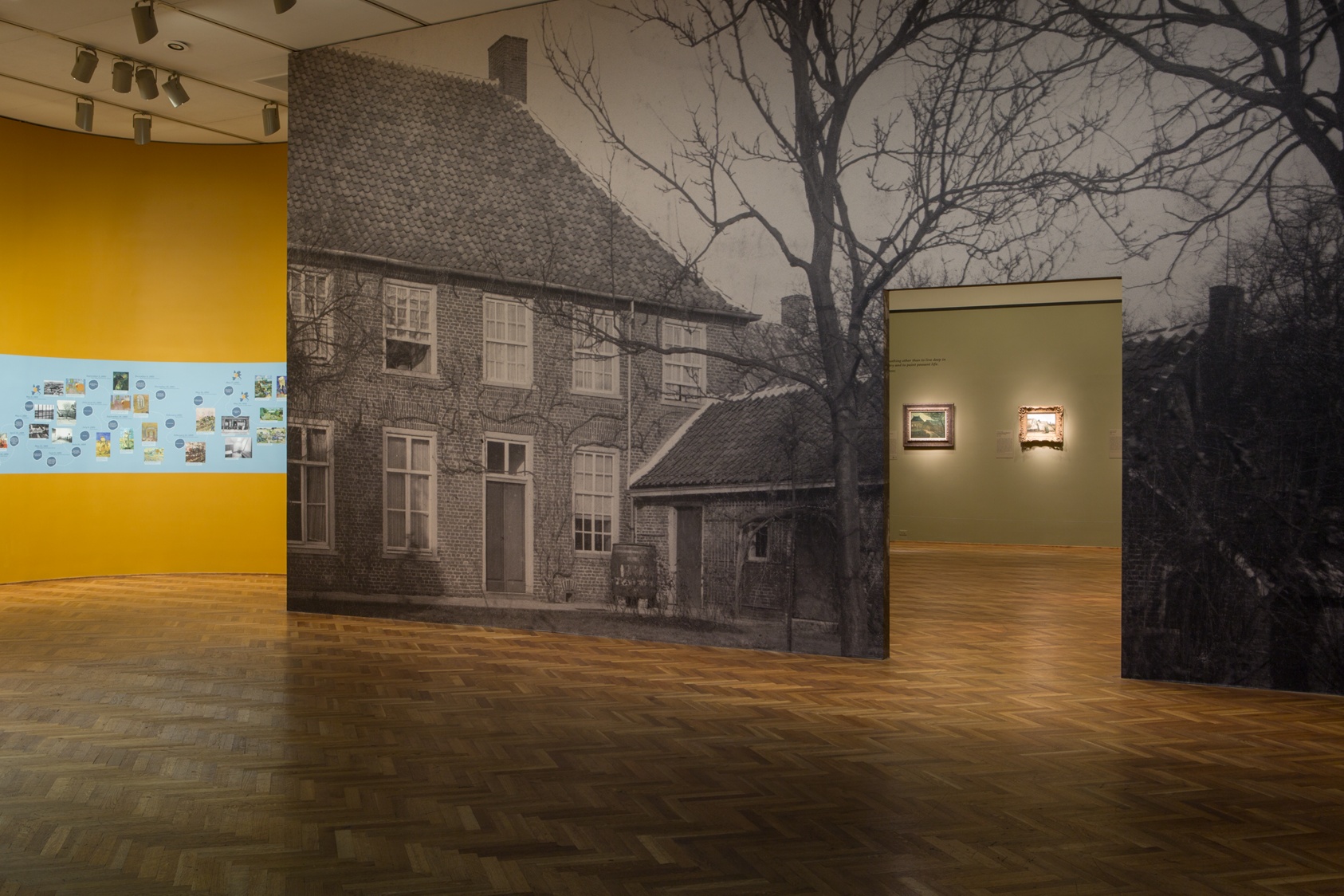 A yellow gallery wall with a blue band containing images is interrupted by a wall jutting into the room depicting a floor-to-ceiling black-and-white image of a two-story home.