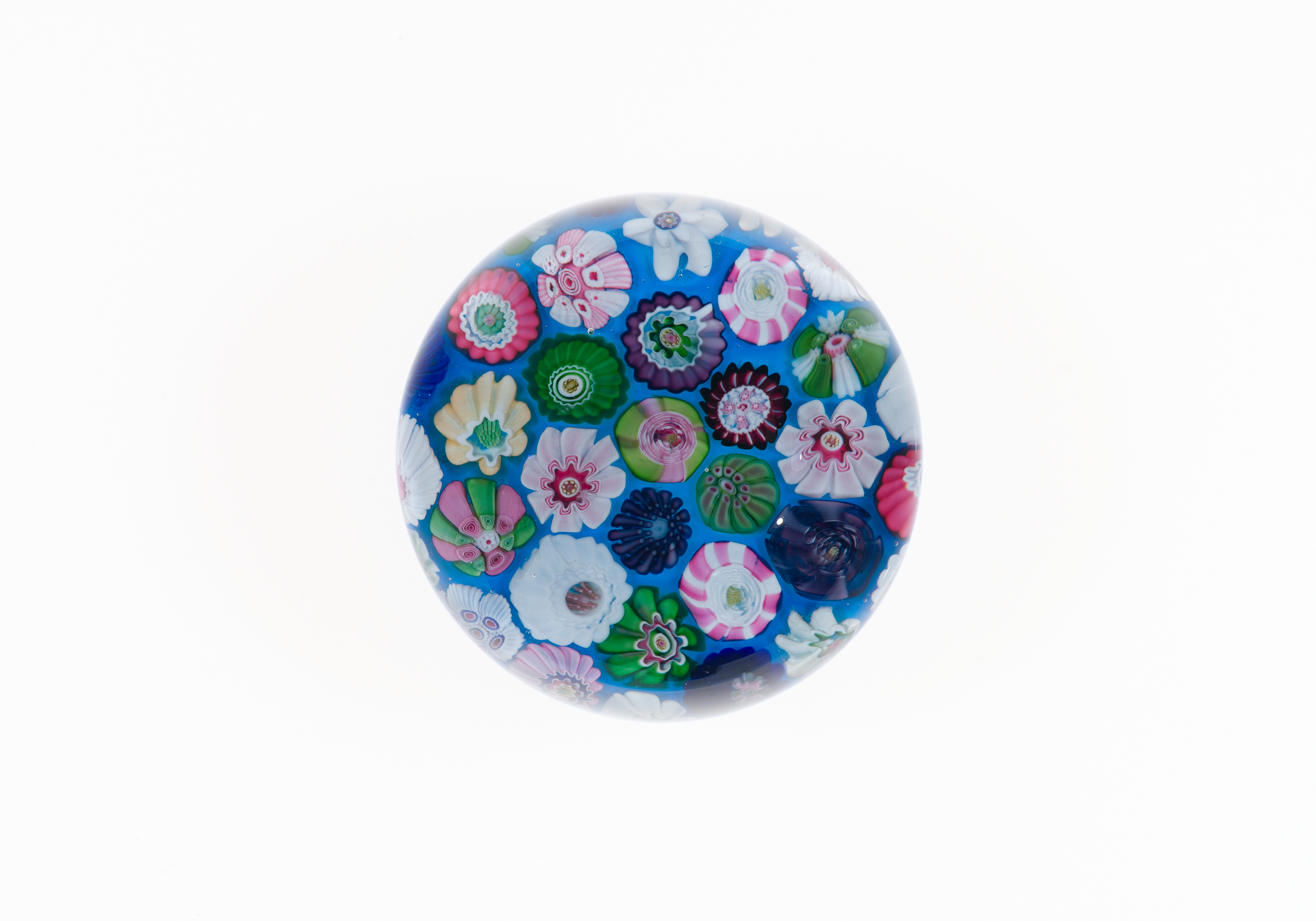 Color photograph of a round glass paperweight at center, its clear dome showing tightly packed flower shapes in pink, green, blue, white, and purple on a deep blue base, with a plain white background around it.
