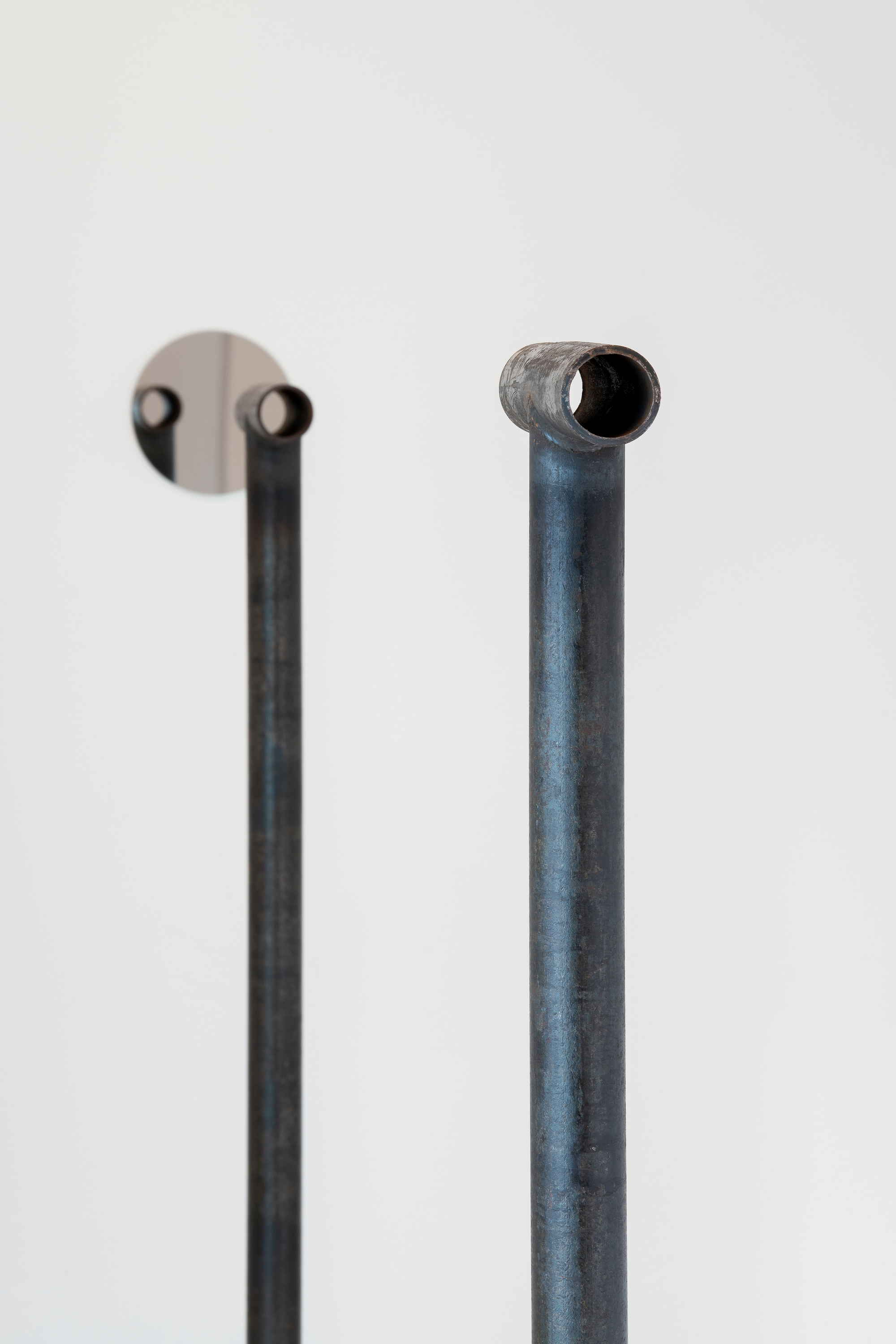 Two black pipes stand vertically with a shorter piece of the same pipe resting on top horizontally. A round mirror on a white wall can be seen through the horizontal pieces of pipe.