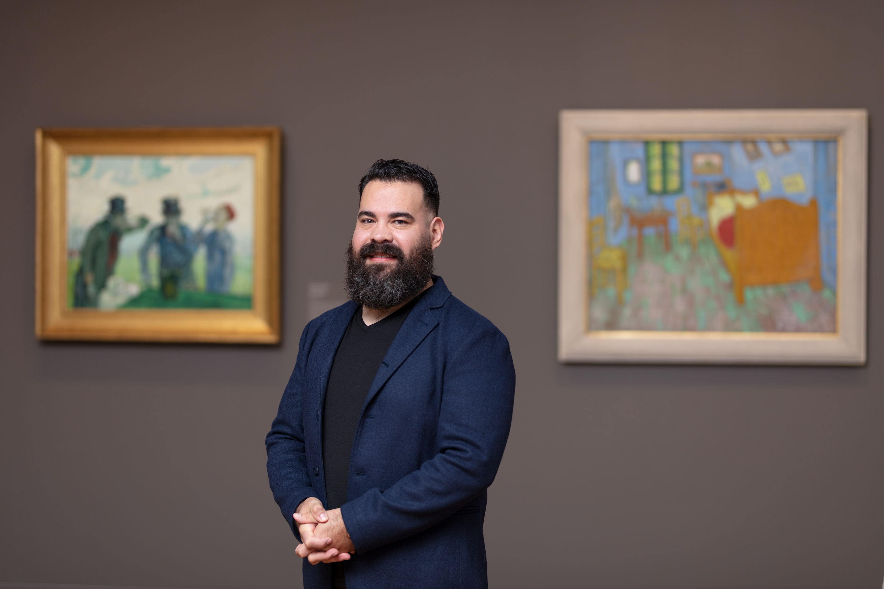 A light-skinned man with dark hair and a full beard, Salvador Cruz, stands between Van Gogh’s “The Drinkers” and “The Bedroom” in a dark-colored art gallery.