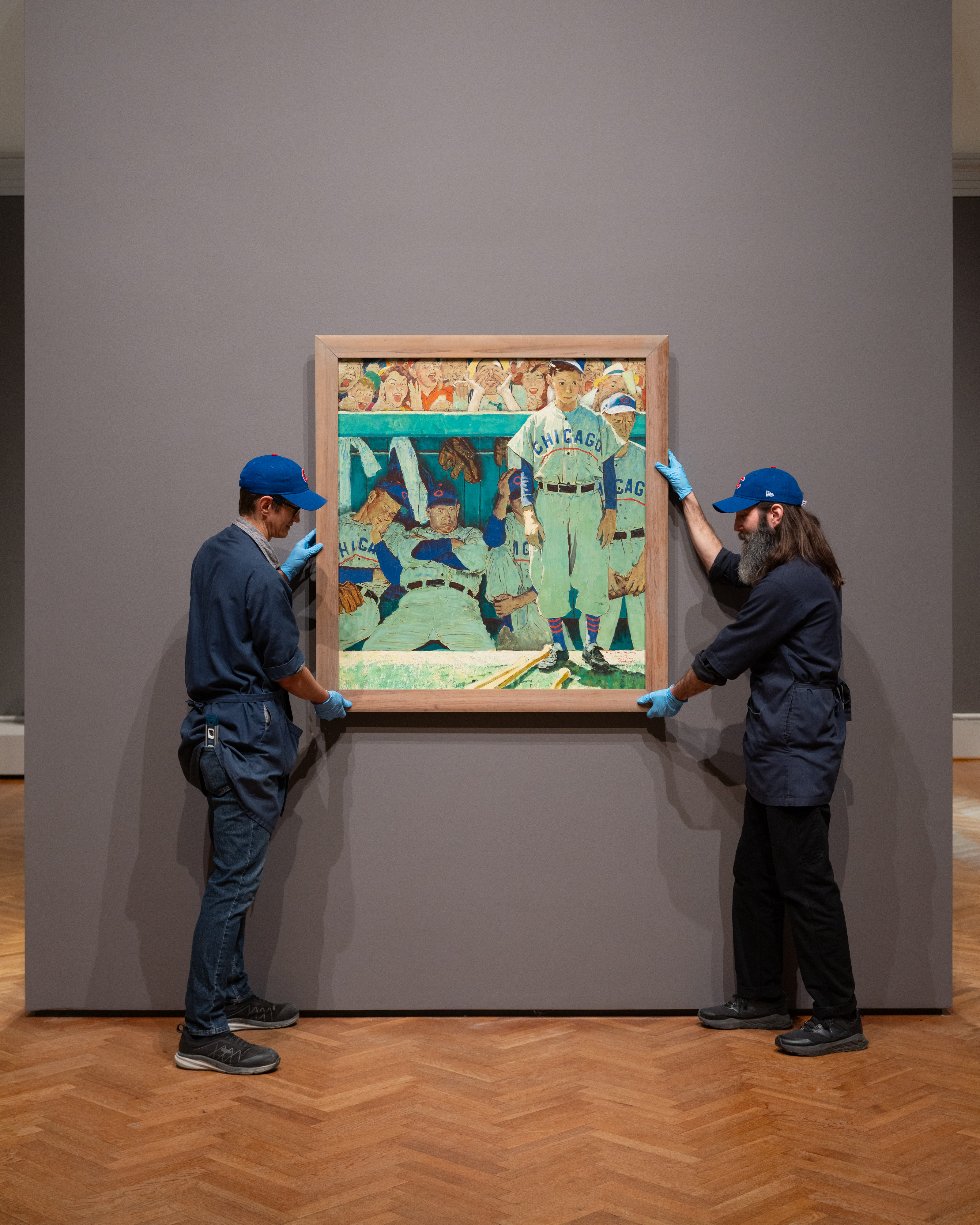 Two men dressed in black and wearing Chicago Cubs hats hang Norman Rockwell's, "The Dugout," on a wall in a museum