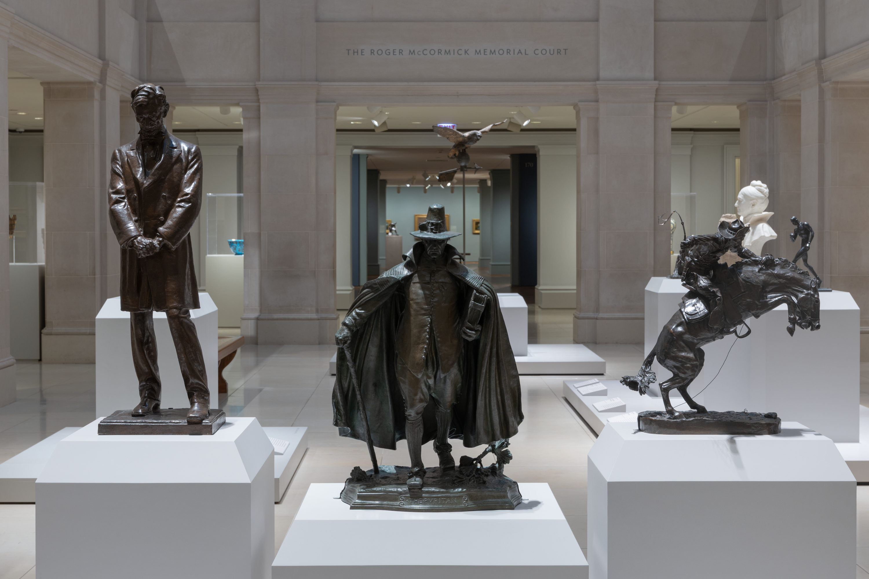 A color photograph shows a museum gallery with three bronze sculptures on white plinths: at left, a standing man in a long coat with hands clasped; at center, a caped figure in a wide-brim hat walking forward; at right, a rider on a rearing horse, with pale stone walls around.