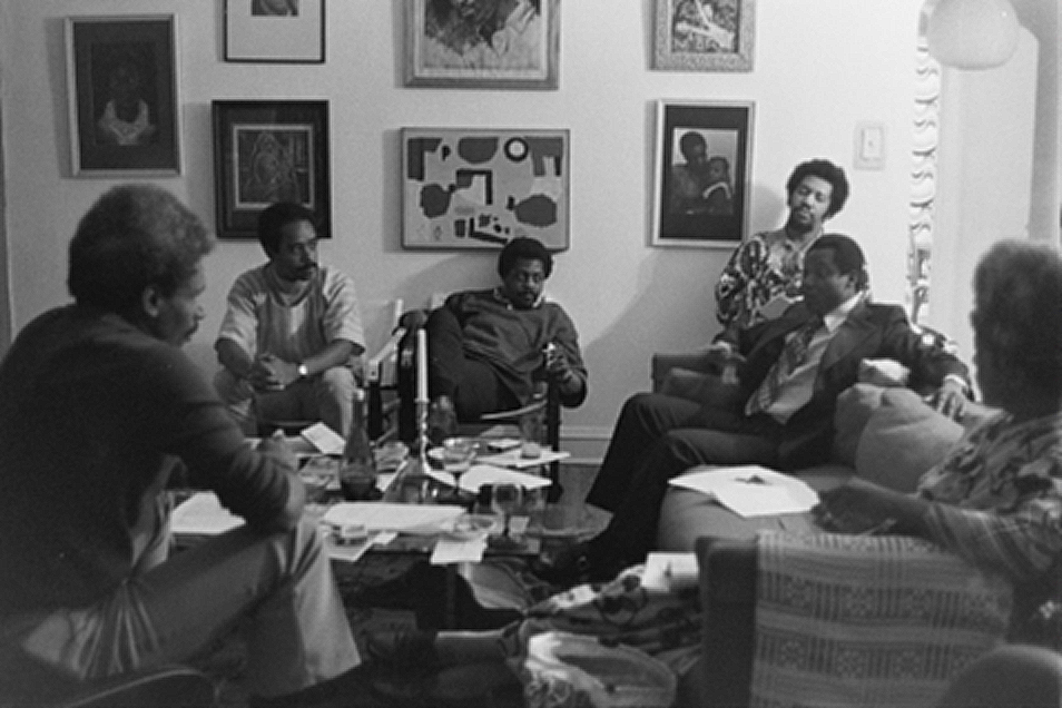 Six people gather in a living room, seated or standing around a coffee table cluttered with papers, notebooks, and bottles. The setting appears informal but focused, with most individuals engaged in discussion. Framed artworks decorate the wall behind them, including abstract and portrait pieces. The group includes men and women, some in suits and others in casual or patterned clothing. A standing woman near the back holds a piece of paper, listening. The lighting is soft, suggesting an intimate planning or strategy meeting.