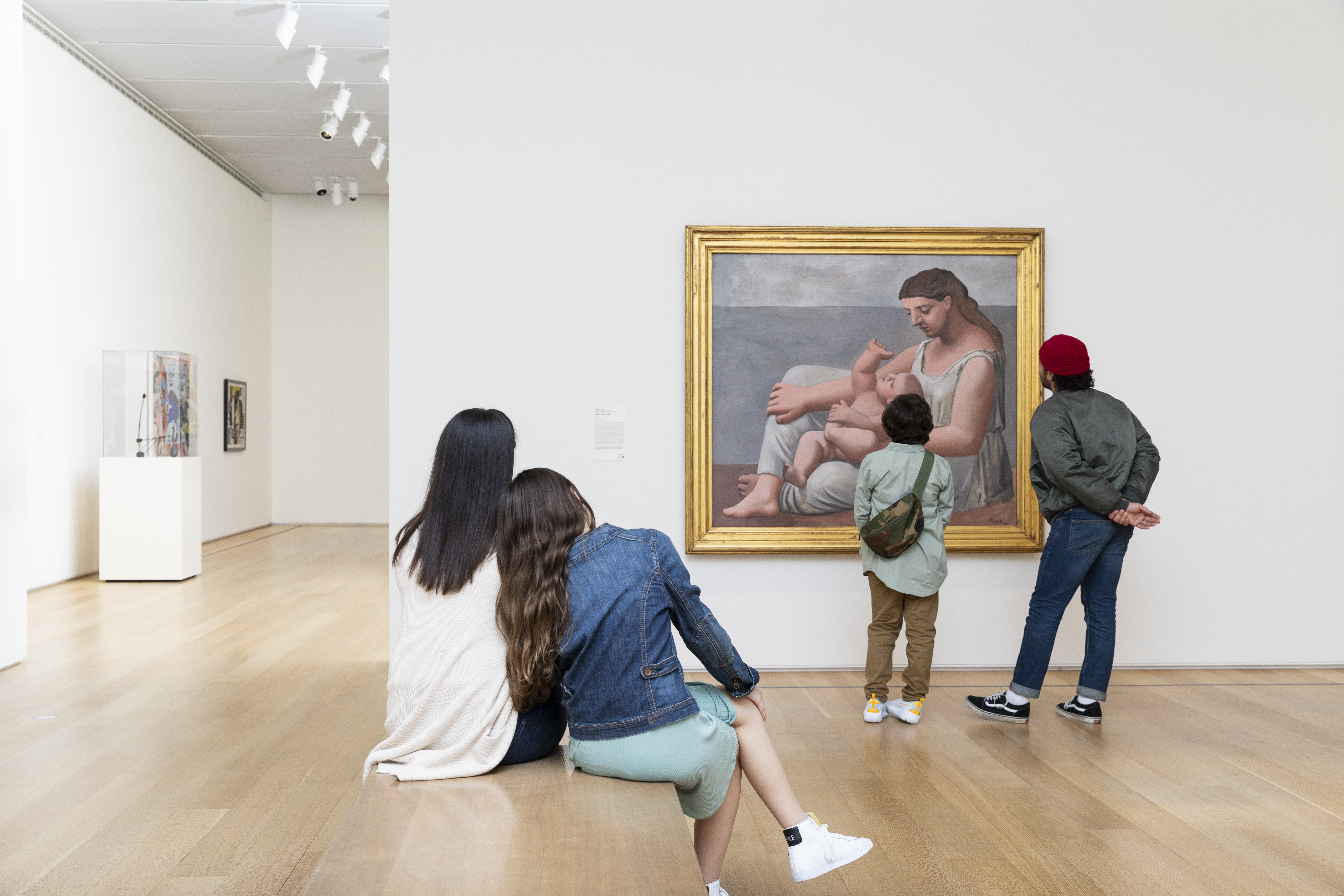 Photograph shows people in an art gallery observing Pablo Picasso's large painting "Mother and Child." On a bench before the painting, a woman and an adolescent girl sit together, the girl's head resting on the woman's shoulder. A young boy gazes up at the painting, his back to us.