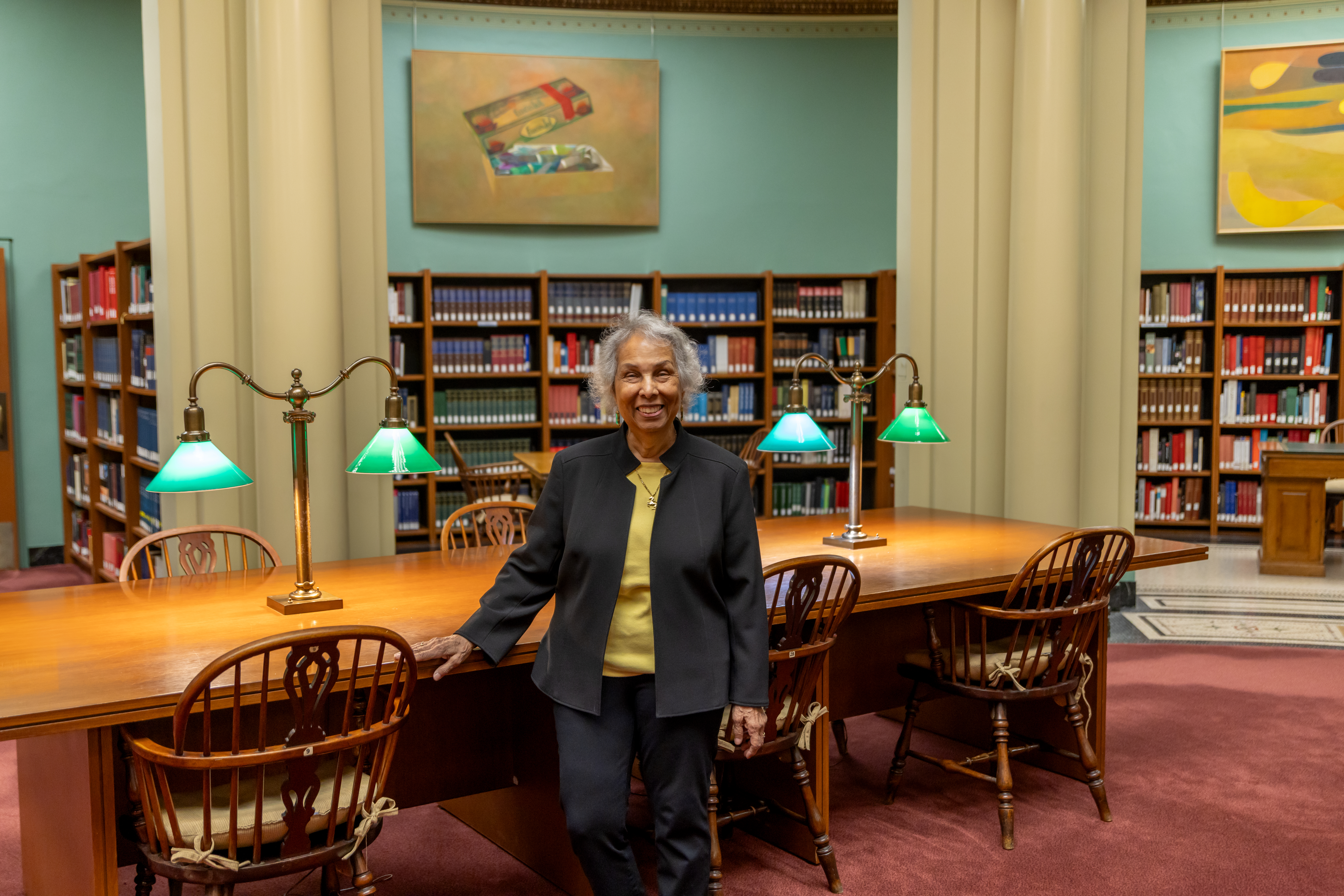Photograph shows Cecile Webster, a Black woman with medium-toned skin, a wide smile, and short gray-white hair, in a dark jacket and yellow shirt. She stands facing the viewer in the Franke Reading Room, a book-lined space with wood shelves, green walls, and large wood tables with brass lamps that have green glass shades.