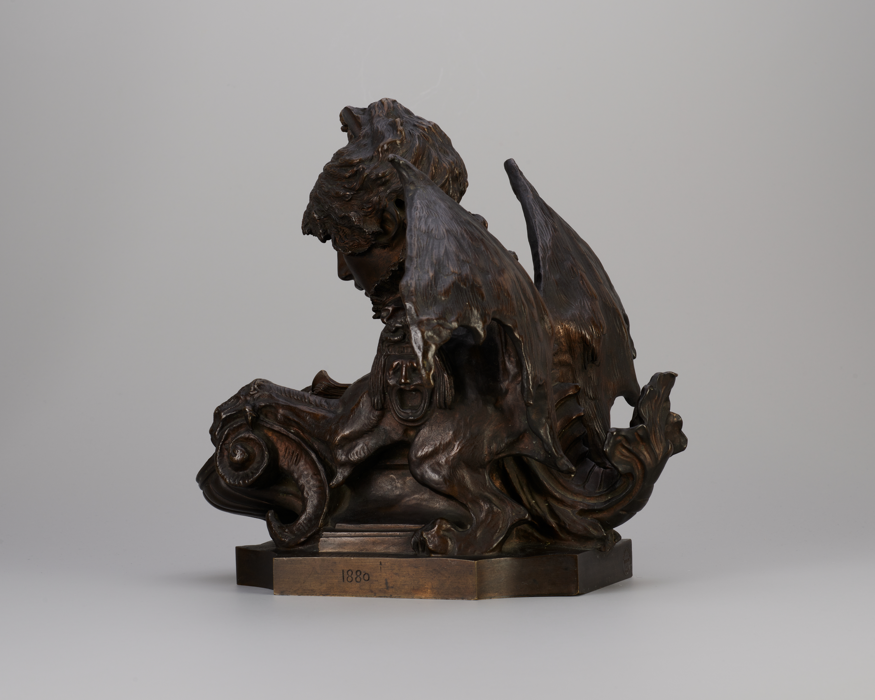 Side view of a a bronze-colored metal sculpture of a creature with the head of a curly-haired young woman, extended bat’s wings, and taloned claws. The figure looks downward and a dragonlike tail curves up at back.