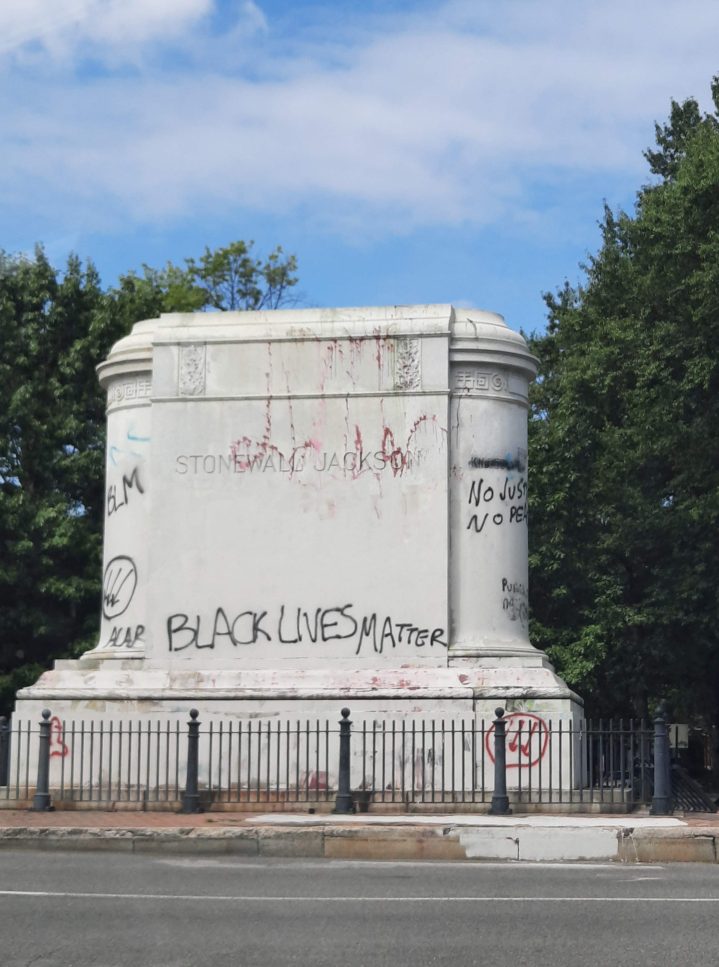 A large empty plinth is covered in graffiti behind an iron fence amid green trees and a bright blue sky with fluffy clouds. One graffito reads BLACK LIVES MATTER. The name STONEWALL JACKSON is carved into the side of the plinth.