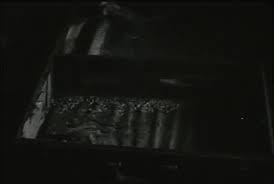 Photograph of a dimly lit interior showing a large, shallow, rectangular container with water inside. Ripples and small debris or particles are visible on the water’s surface. Diagonal light shines across the container, casting strong shadows. The setting is dark and industrial in tone, with only partial details visible.