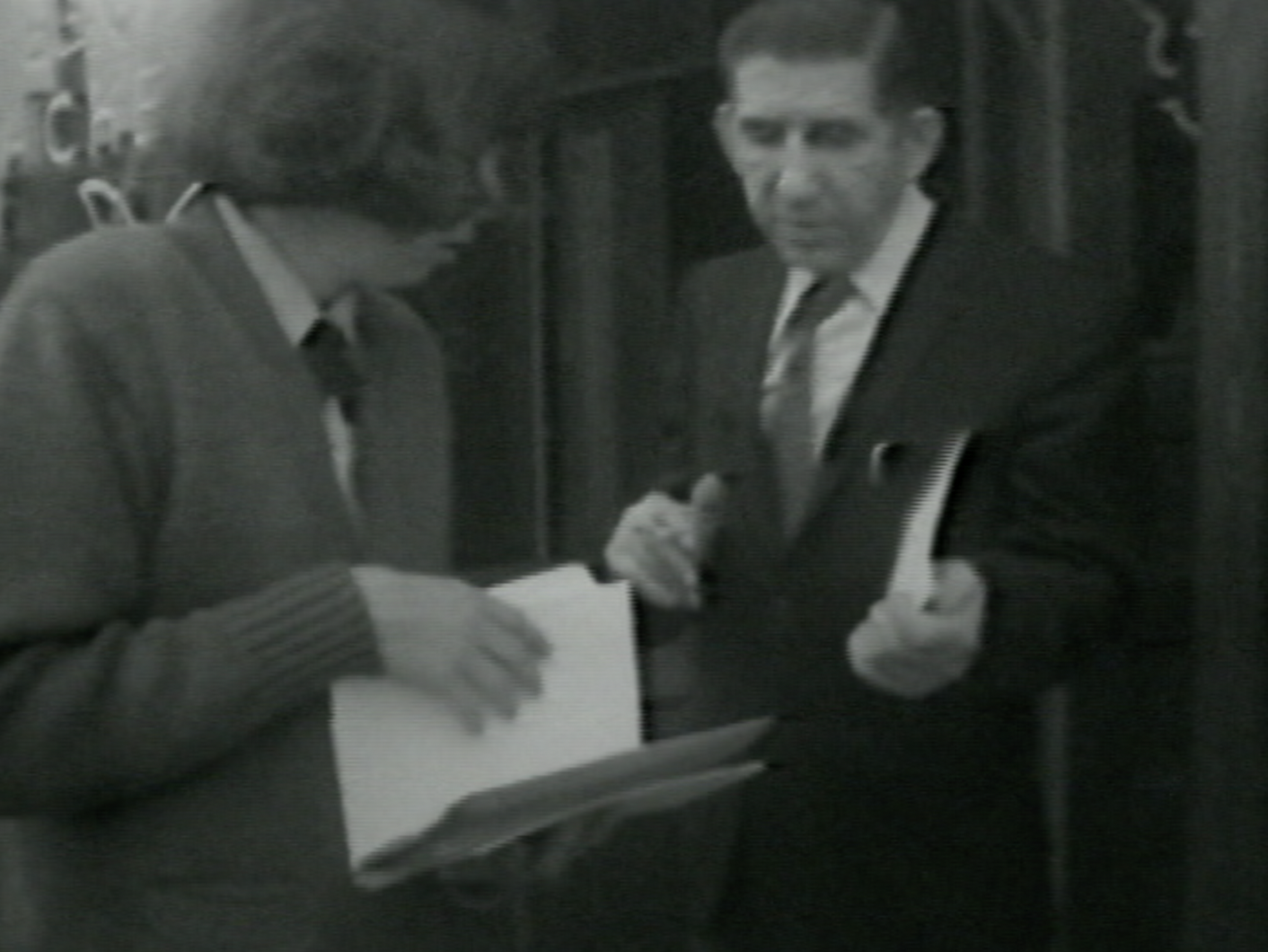 Black-and-white photograph of two people standing near a doorway. On the left, a person with shoulder-length hair and a bulky sweater holds a clipboard and large envelope. On the right, a man in a suit and tie holds a piece of mail, facing the other person while speaking. Both are engaged in conversation, with the doorway and partially visible signage behind them.