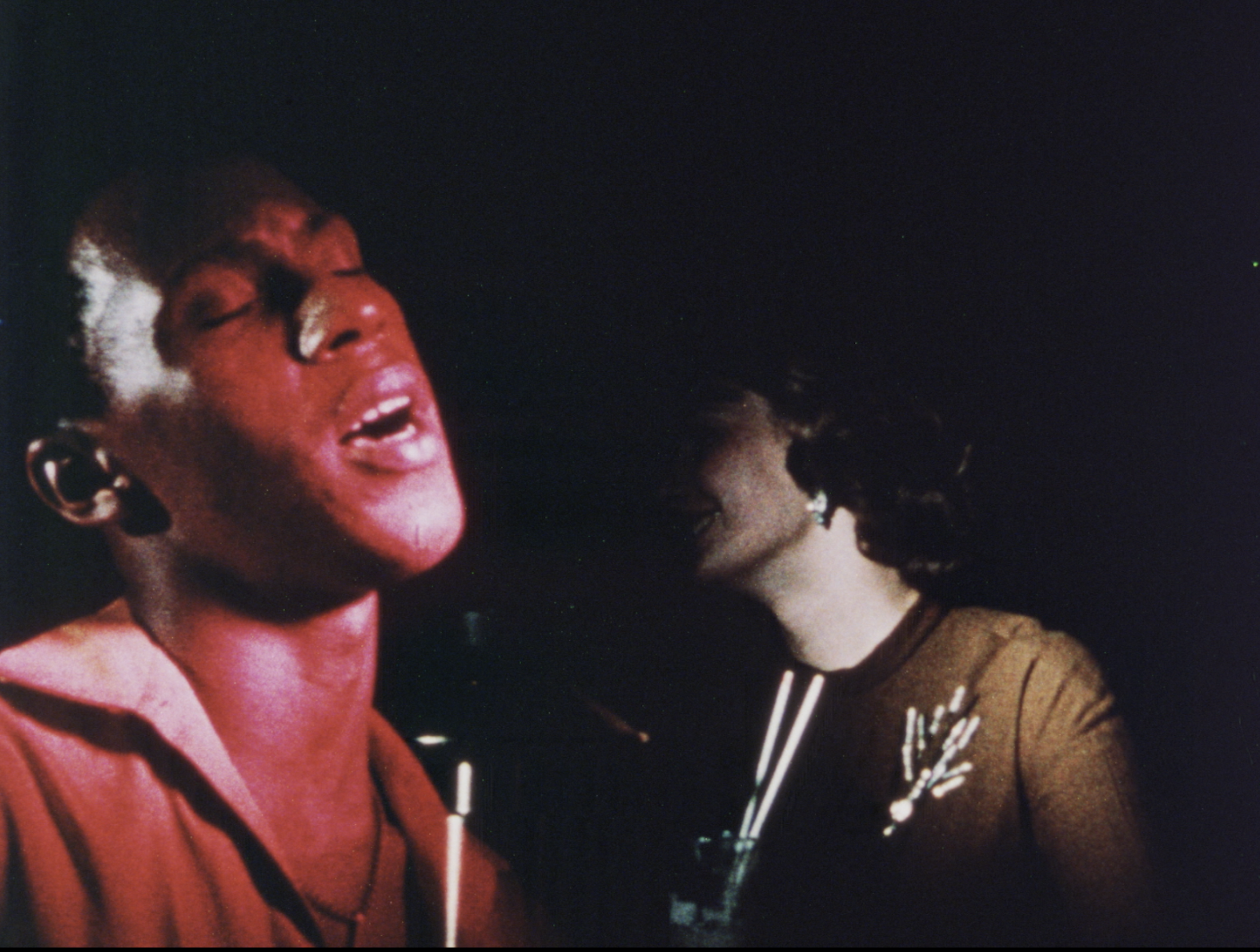 Close-up of a man singing with eyes closed, his face lit with deep red lighting, suggesting stage lights or a nightclub setting. He wears a red open-collared shirt and leans toward a microphone at the bottom center. Behind him in shadow, a woman in a brown dress with a rhinestone brooch and earrings smiles, holding a drink with two white straws. The background is dark, emphasizing the singer’s expressive face.