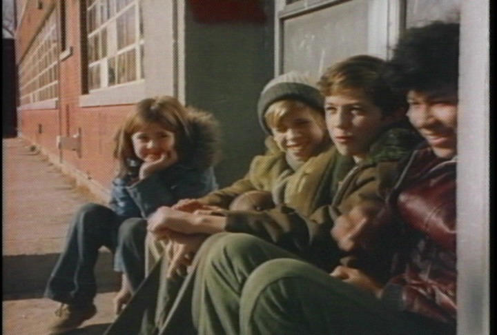 Four children sit on a concrete stoop outside a building with red brick and large windows. All are dressed for cold weather in puffy jackets, hats, and scarves. The child on the far left leans on one hand and smiles directly at the camera. The others sit close together, laughing and looking toward something out of frame to the right. The afternoon sunlight casts shadows across the sidewalk.