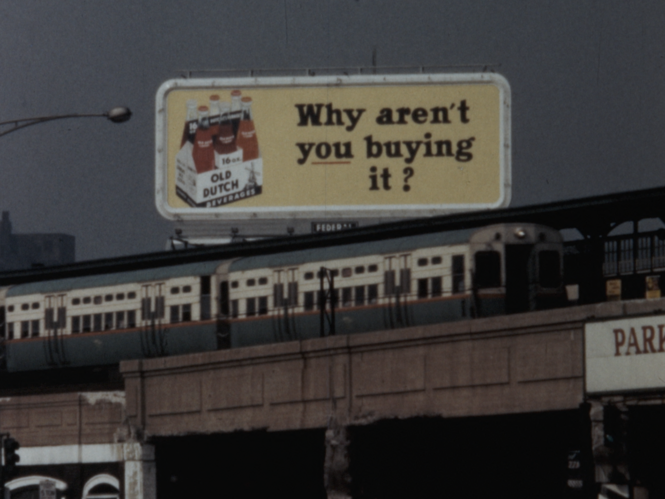 Photograph of an elevated train passing beneath a billboard advertising Old Dutch Beverages. The billboard, set against a hazy gray sky, shows a six-pack of 16 oz glass bottles with red labels and white caps. Large black and red text on a yellow background reads, “Why aren’t you buying it?” The train below is silver with a teal and orange stripe and is partially obscured by the billboard’s support structure. The lower portion of the frame includes a sign reading “PARK” in red capital letters on a white background.